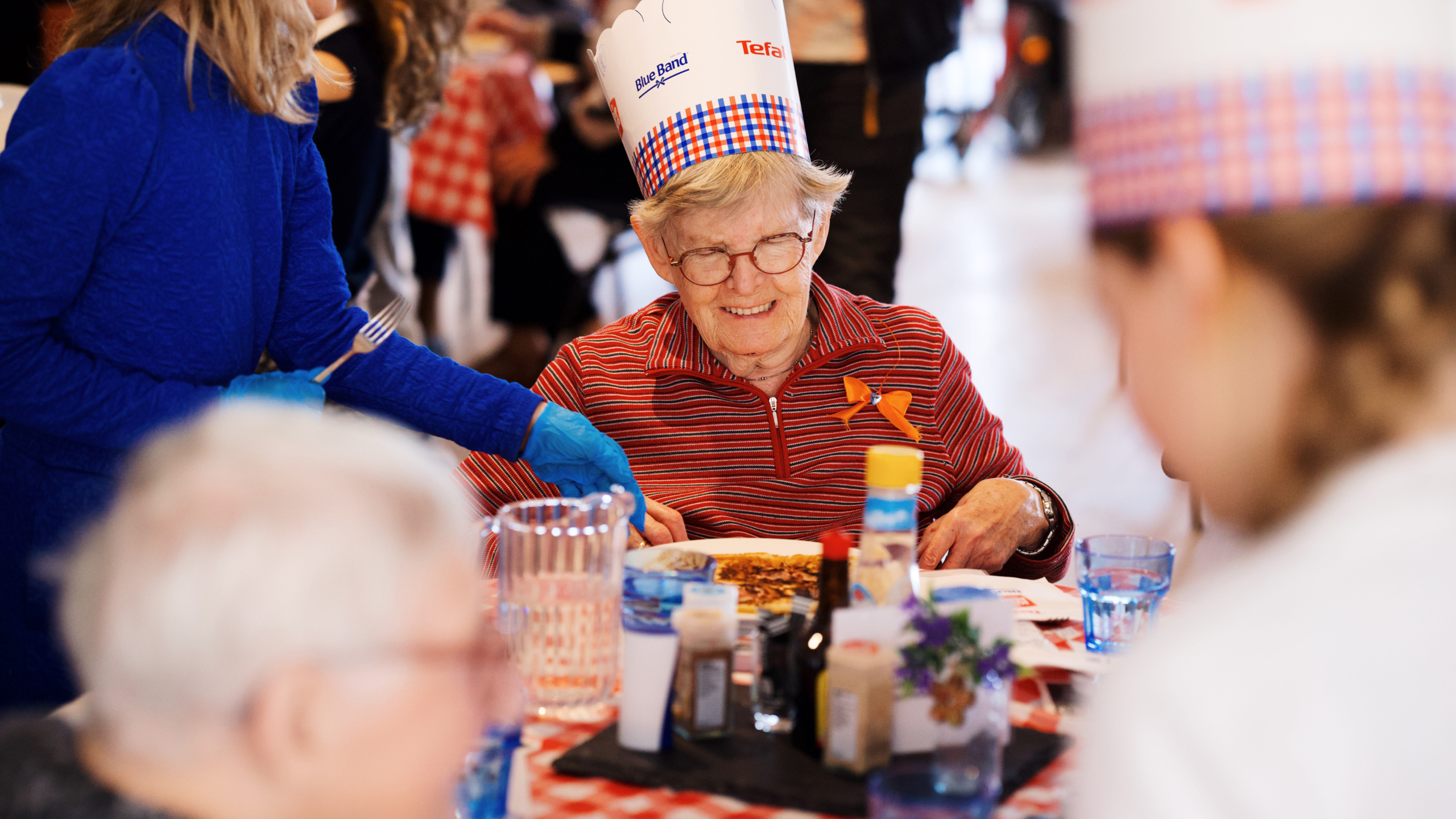 Een oudere vrouw met een papieren koksmuts eet aan een feestelijk gedekte tafel, omringd door andere mensen.
