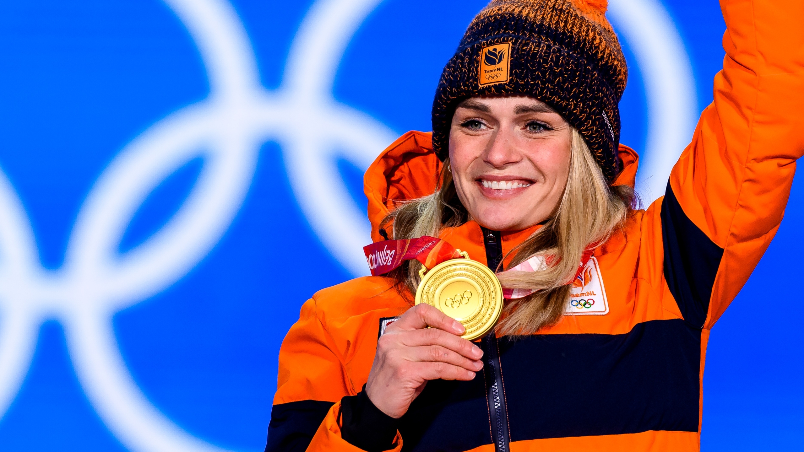 Atlete draagt een oranje jas en muts, houdt gouden medaille vast en glimlacht op podium met Olympische ringen op achtergrond.