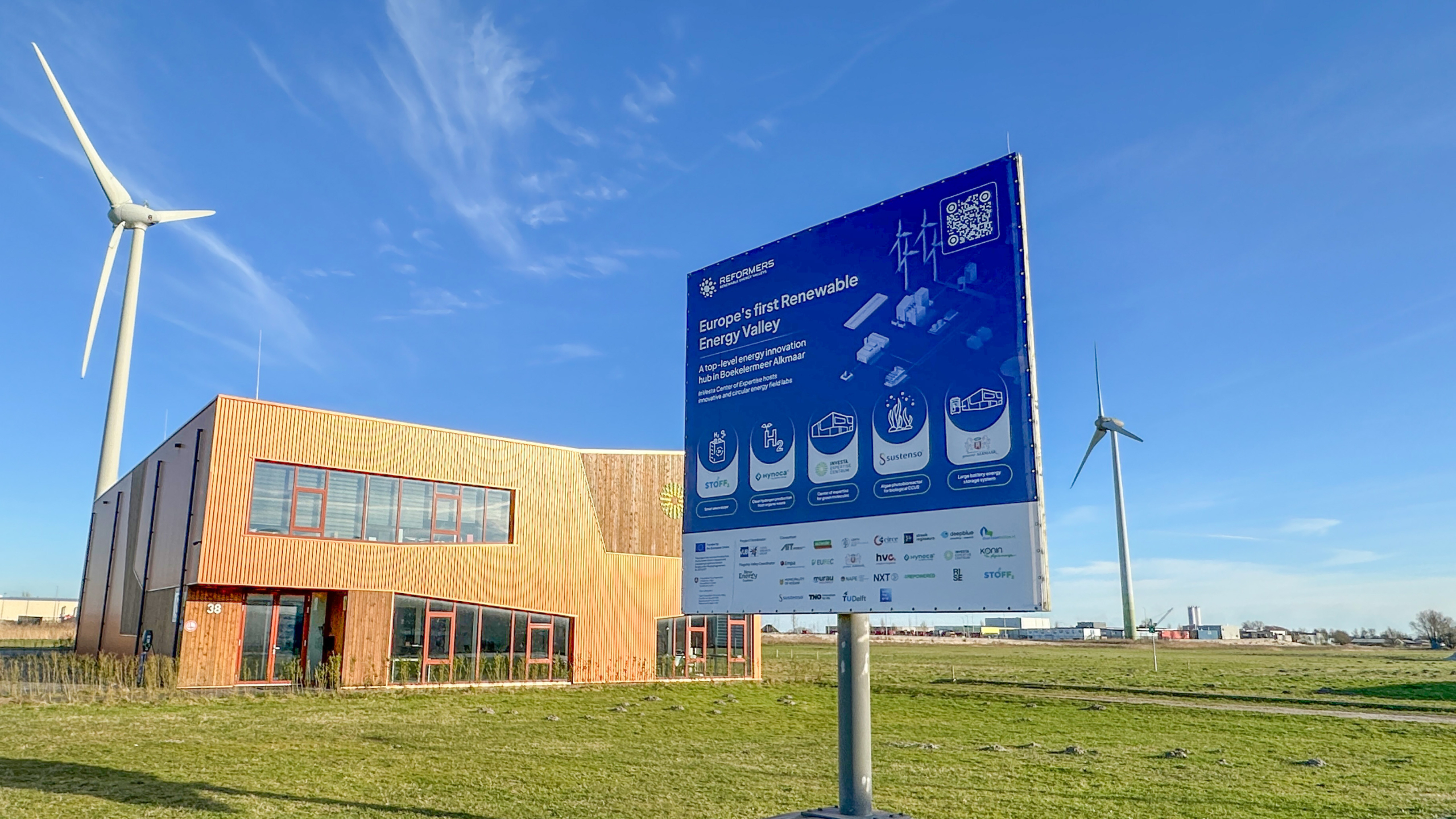 Bord met tekst over Europa's eerste Renewable Energy Valley bij een houten gebouw en twee windturbines in een graslandschap.