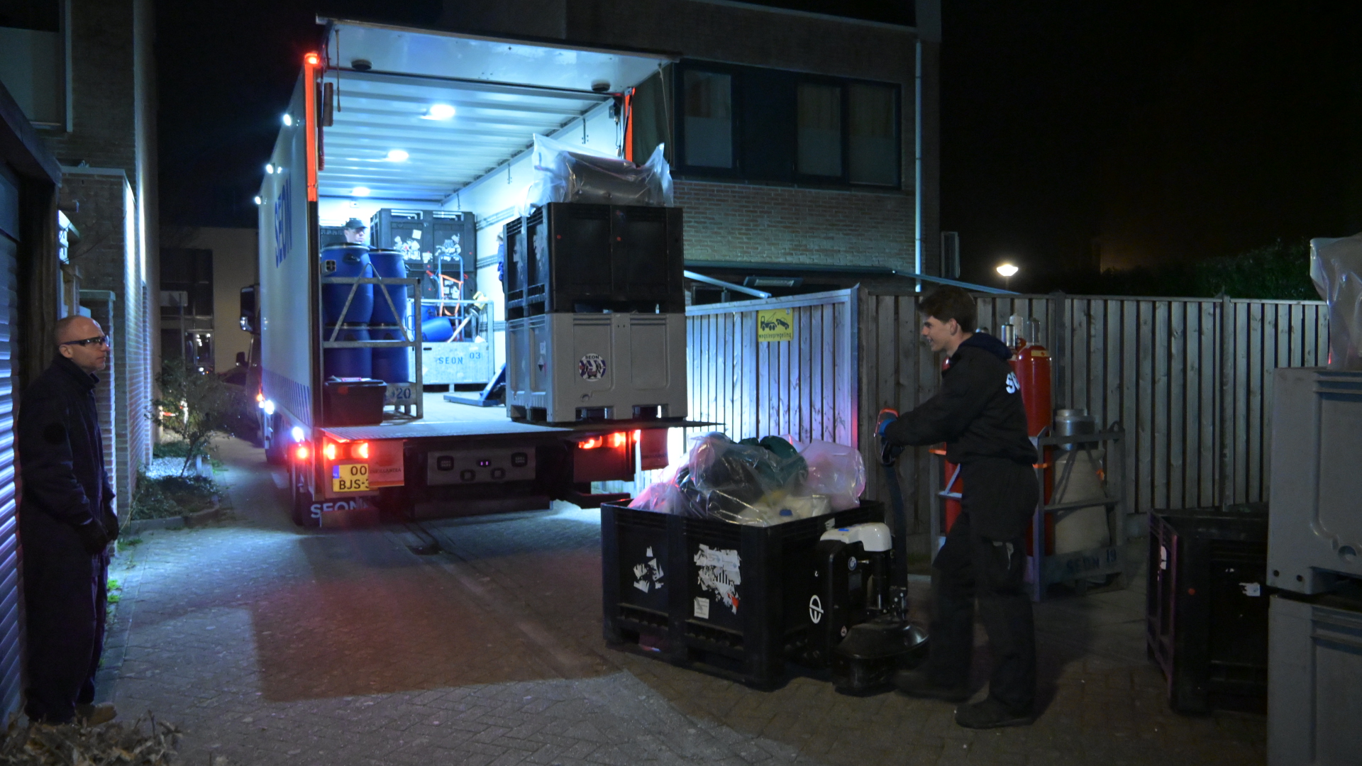 Twee mannen laden 's nachts een vrachtwagen uit met behulp van kratten en vaten in een smalle straat.