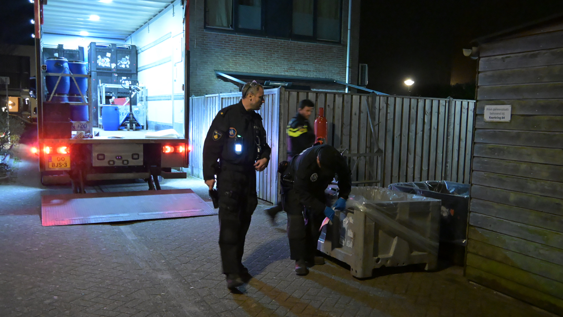 Politieagenten onderzoeken 's nachts een container bij een geopende vrachtwagen op een rustige straat.