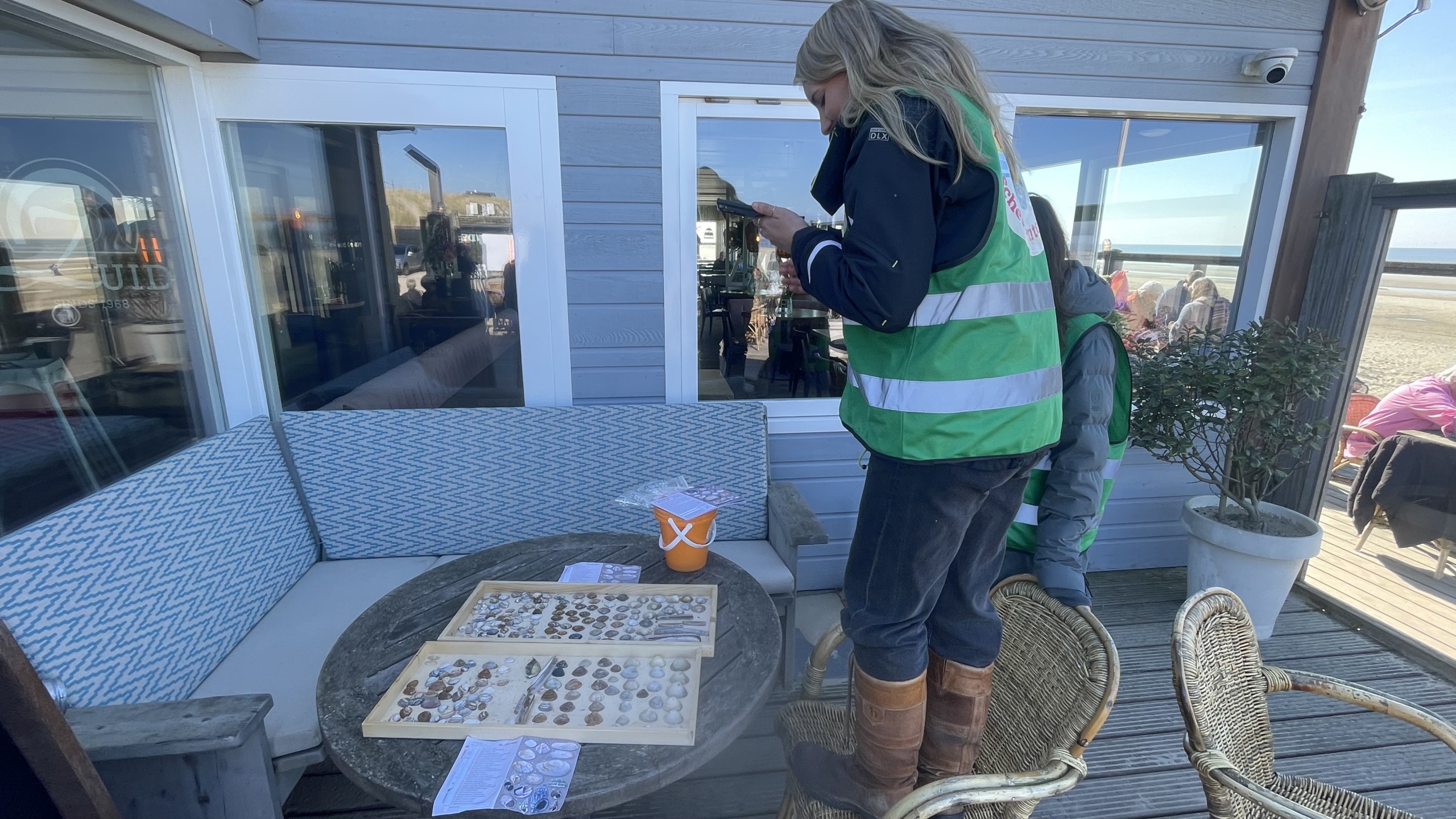 Een persoon in een groene veiligheidshes fotografeert schelpen op een tafel op een terras bij een gebouw aan het strand.