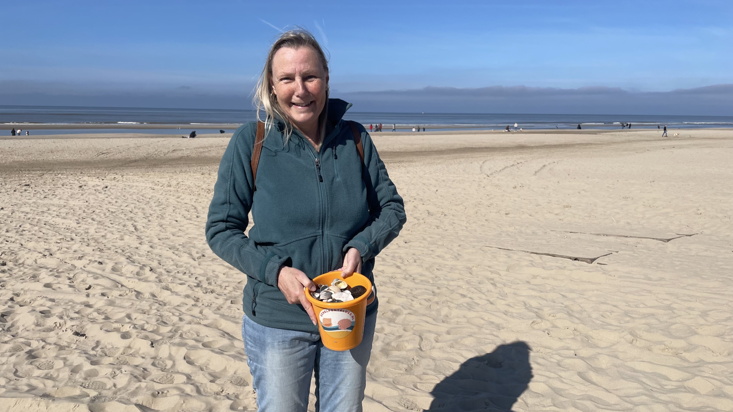 Vrouw glimlacht op een strand terwijl ze een oranje emmer met schelpen vasthoudt.