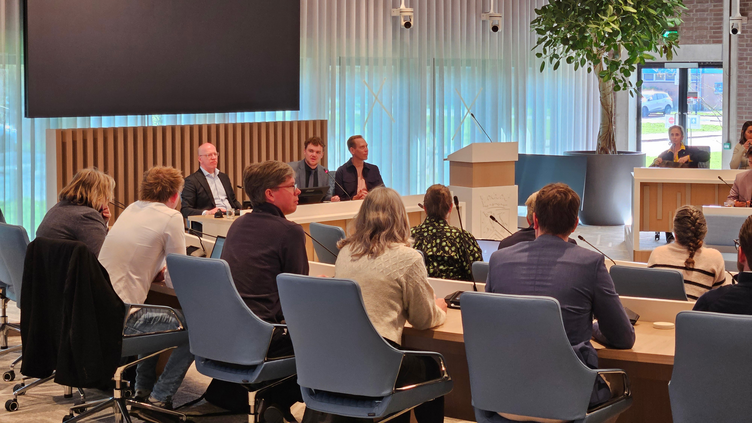 Vergaderruimte met mensen die rond een ovale tafel zitten; een man spreekt terwijl anderen luisteren.