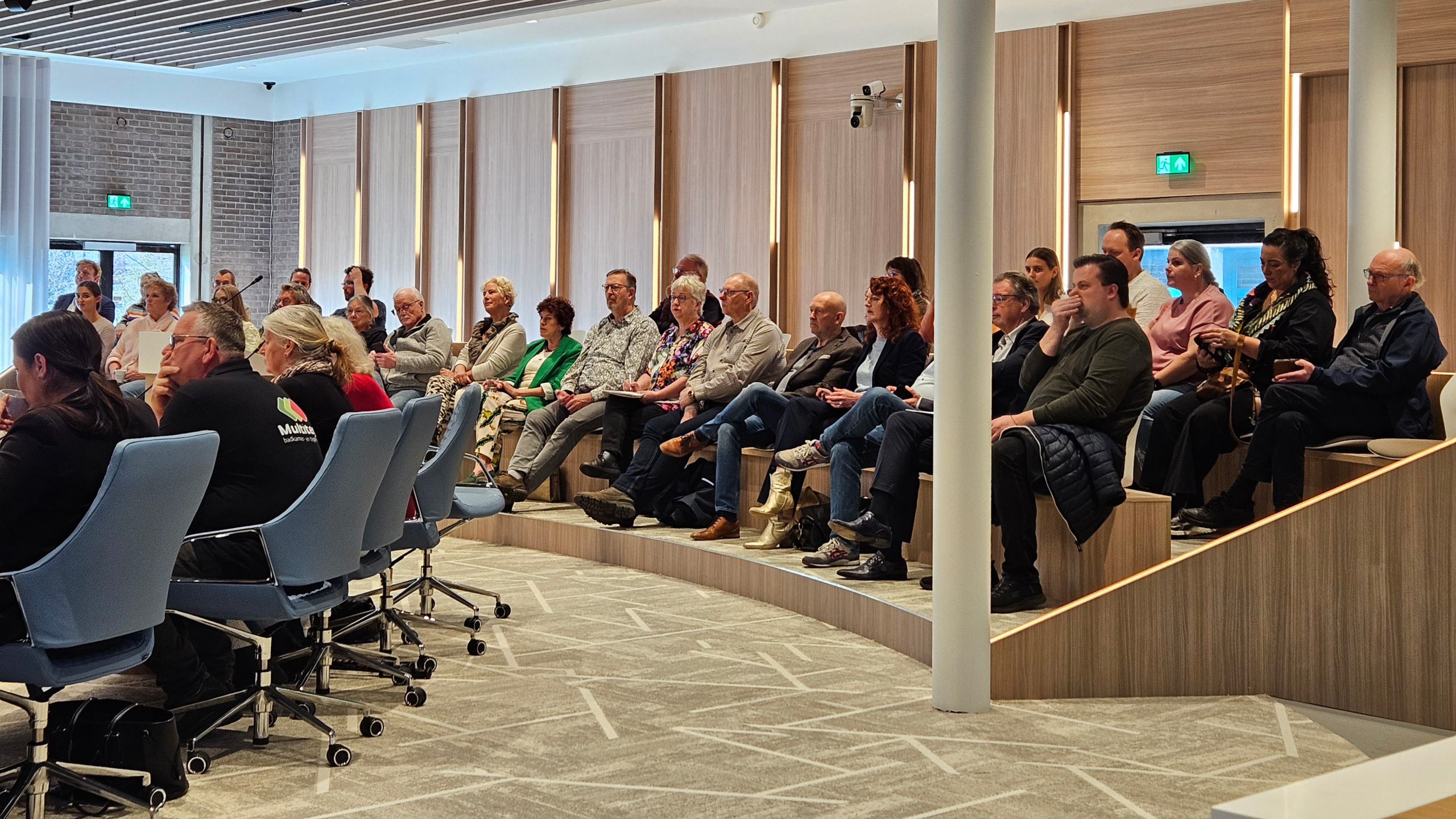 Mensen zitten in een moderne vergaderruimte met gestoffeerde stoelen en een houten afwerking, aandachtig naar een spreker luisterend.