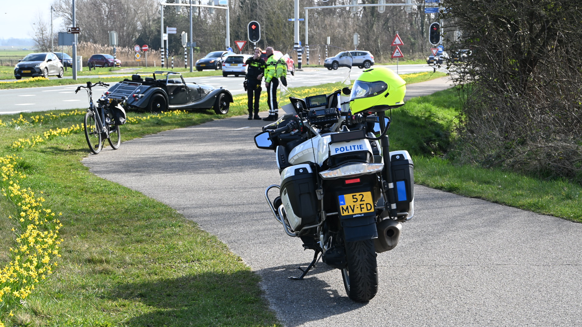 Politiemotor op een fietspad naast een klassieke auto en een fiets, met agenten op de achtergrond in gesprek.