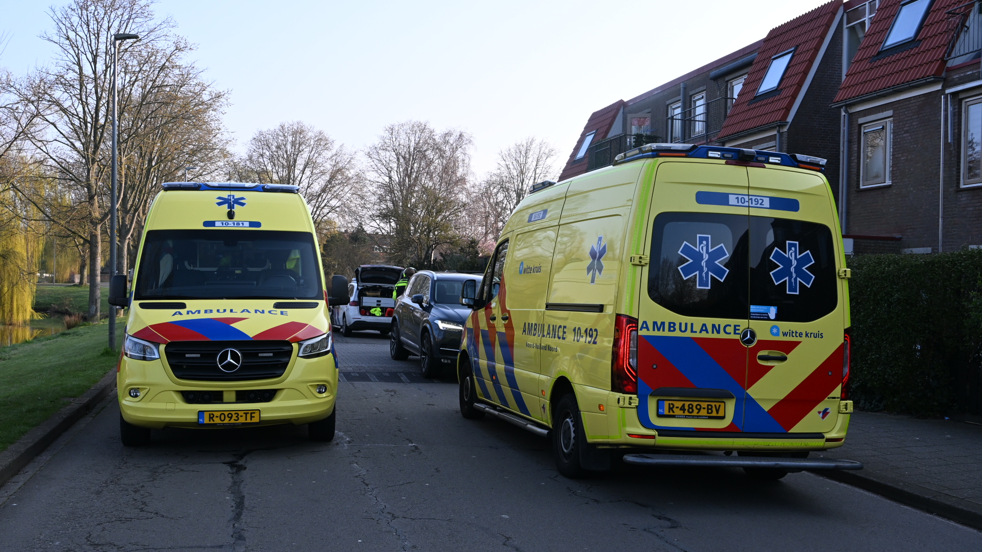 Twee gele ambulances met rode en blauwe strepen staan geparkeerd in een woonstraat, omringd door bomen en huizen.