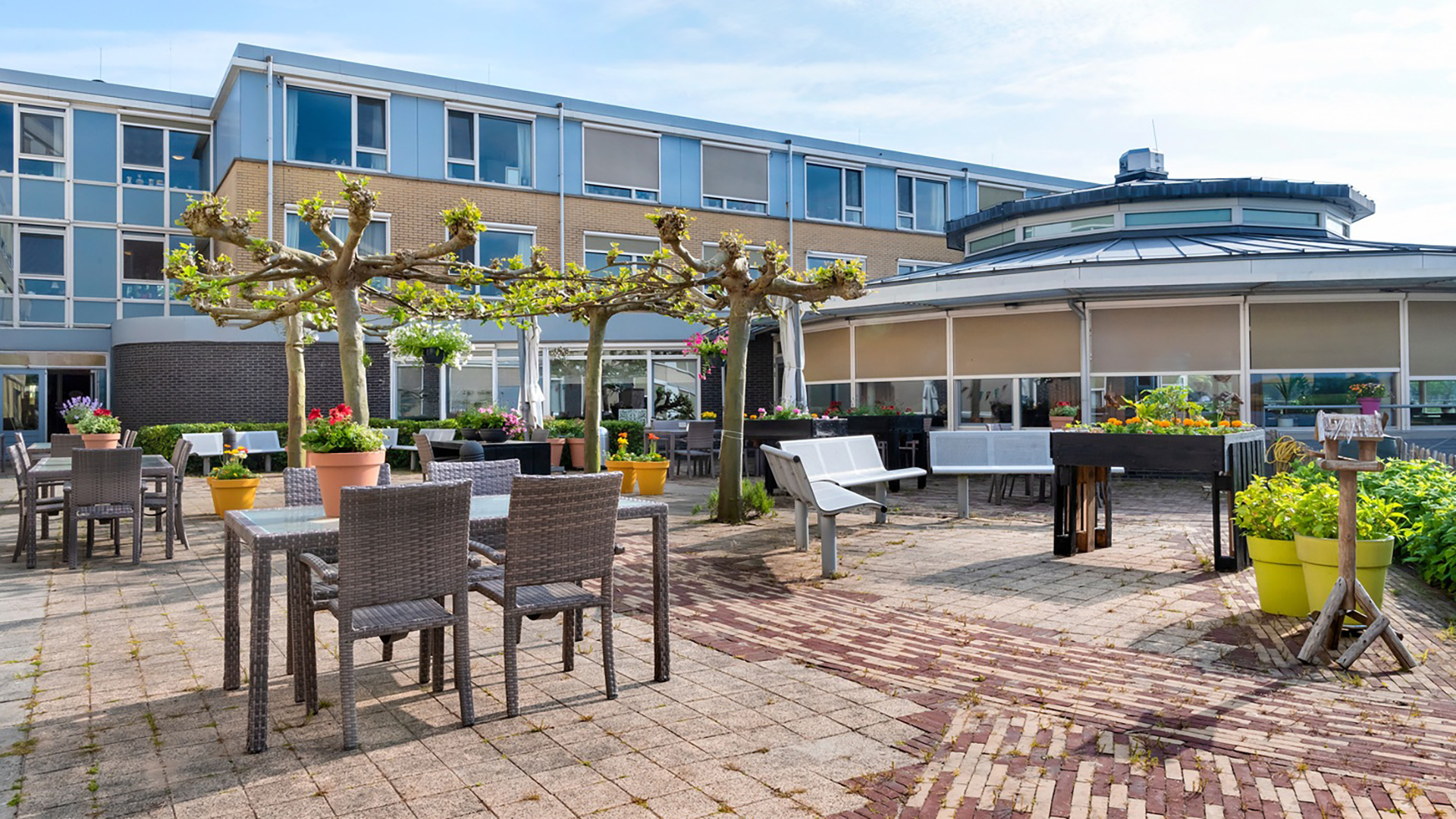 Buitenplaats met tafels, stoelen en plantenbakken omringd door een gebouw met grote ramen.