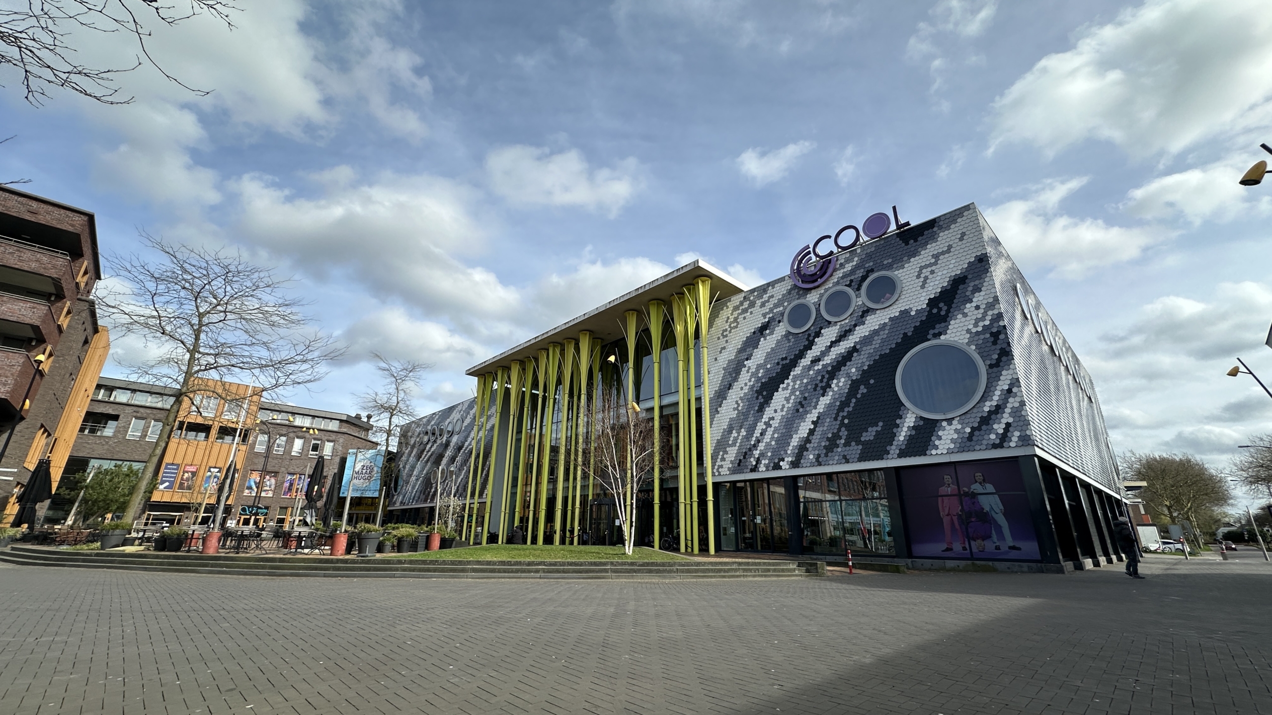 Modern gebouw met opvallende gele zuilen en een mozaïekachtige gevel in een stedelijke omgeving onder een blauwe lucht.