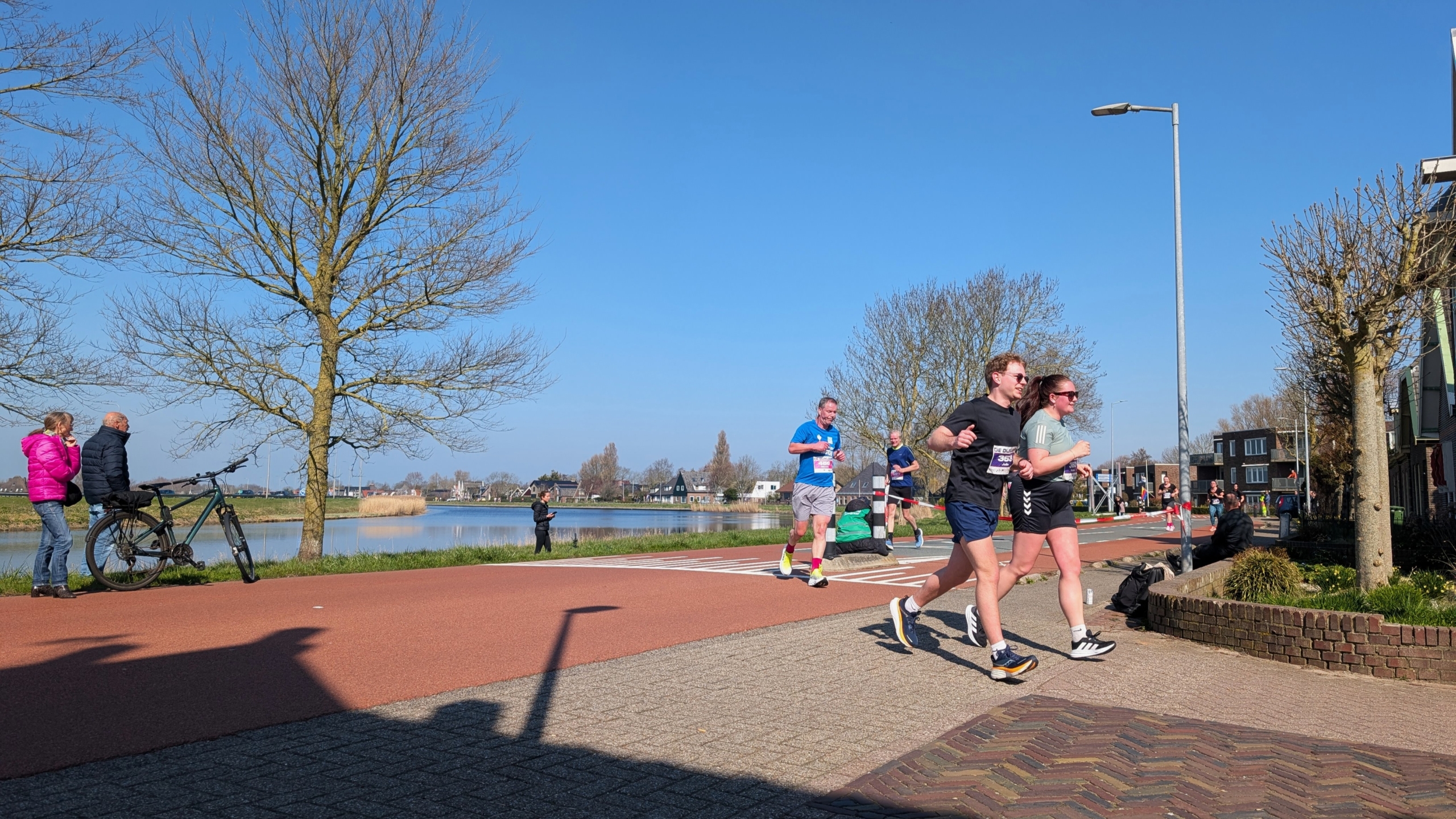 Mensen rennen op een zonnige dag langs een waterloop; toeschouwers staan bij een boom en een fiets.