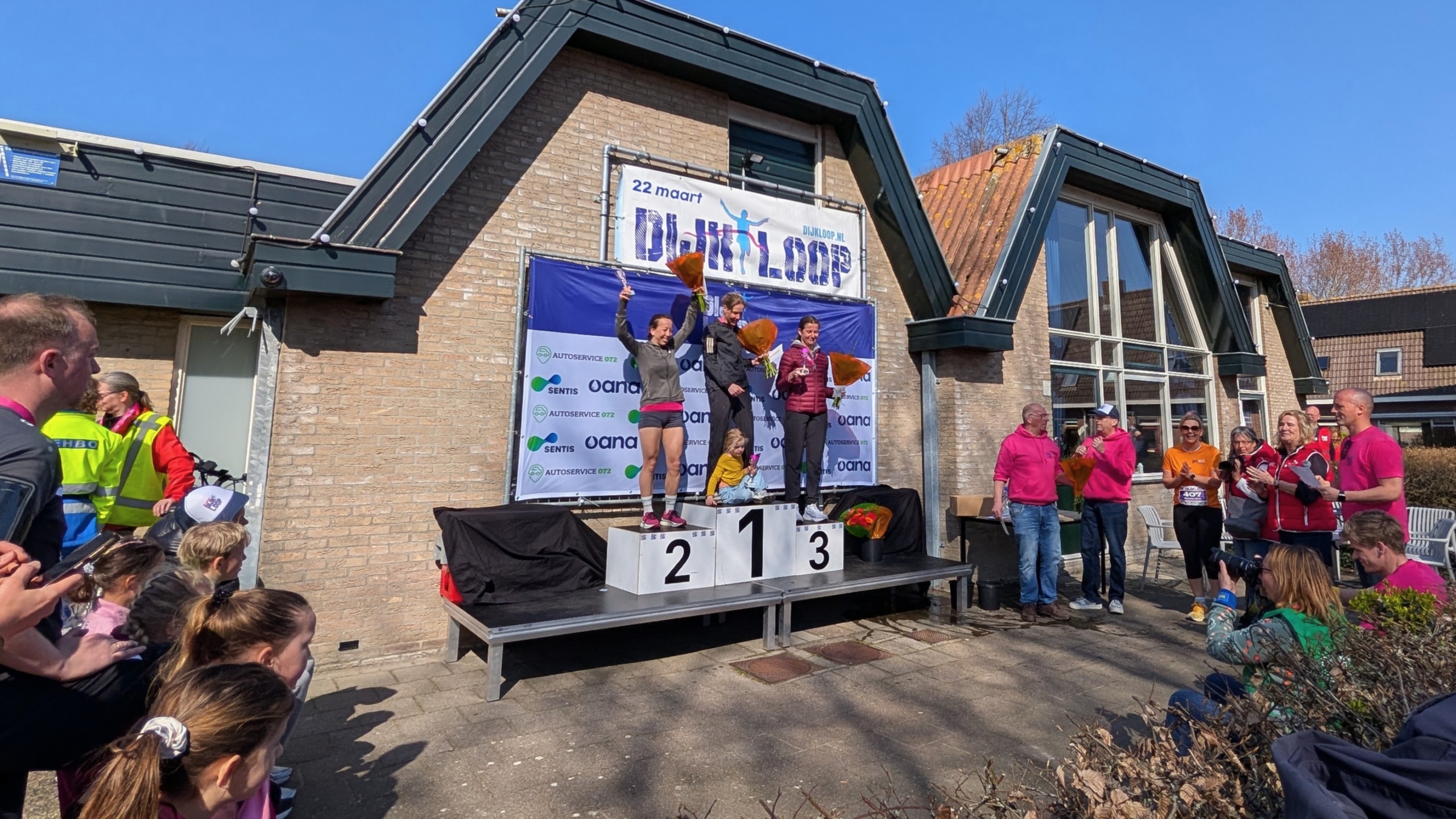 Drie vrouwen op een podium met bloemen tijdens een hardloopevenement voor een publiek, met een groot spandoek op de achtergrond.