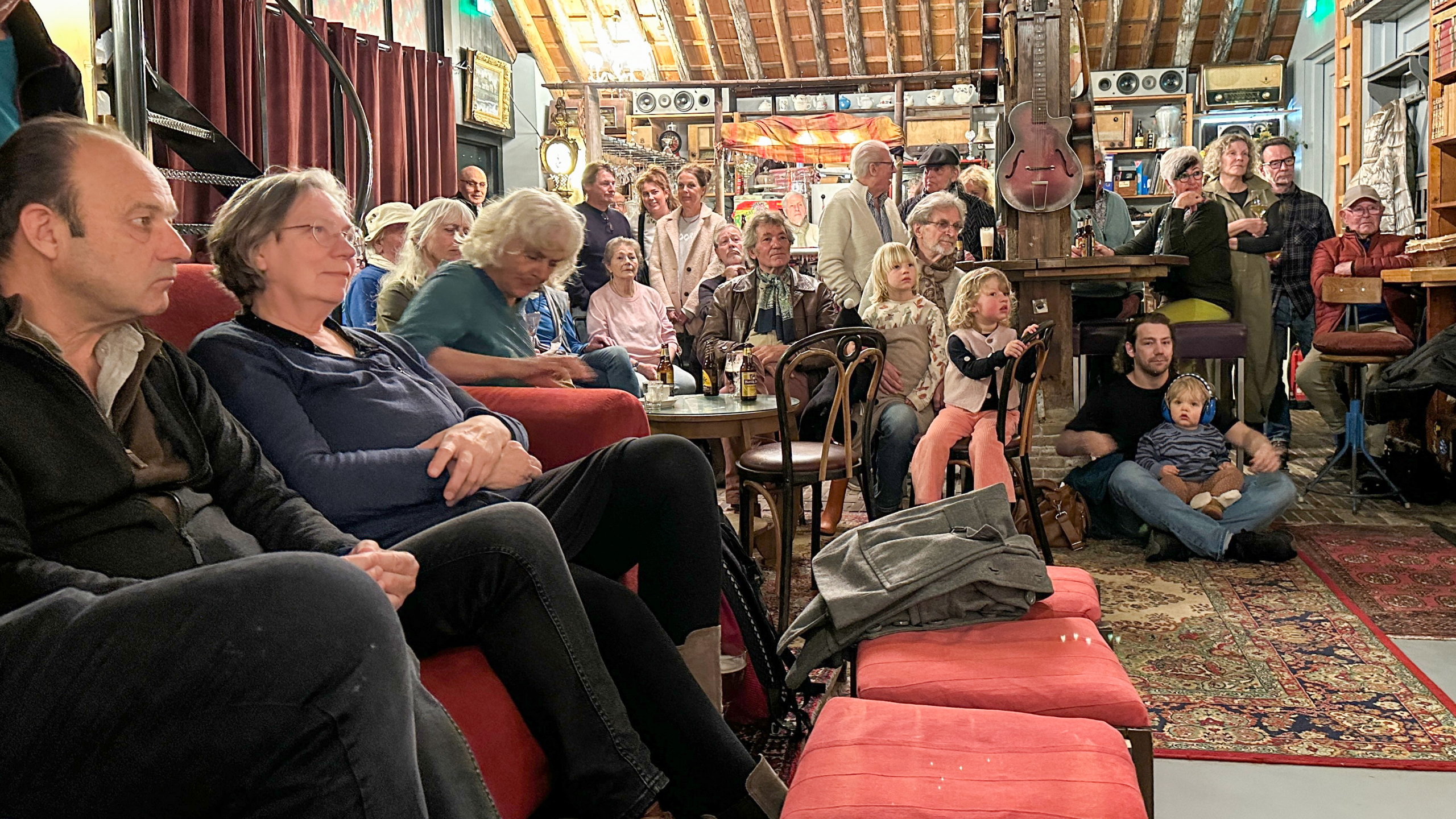 Een groep mensen van verschillende leeftijden zit en staat in een ruimte met een houten plafond, aandachtig kijkend naar iets buiten beeld.