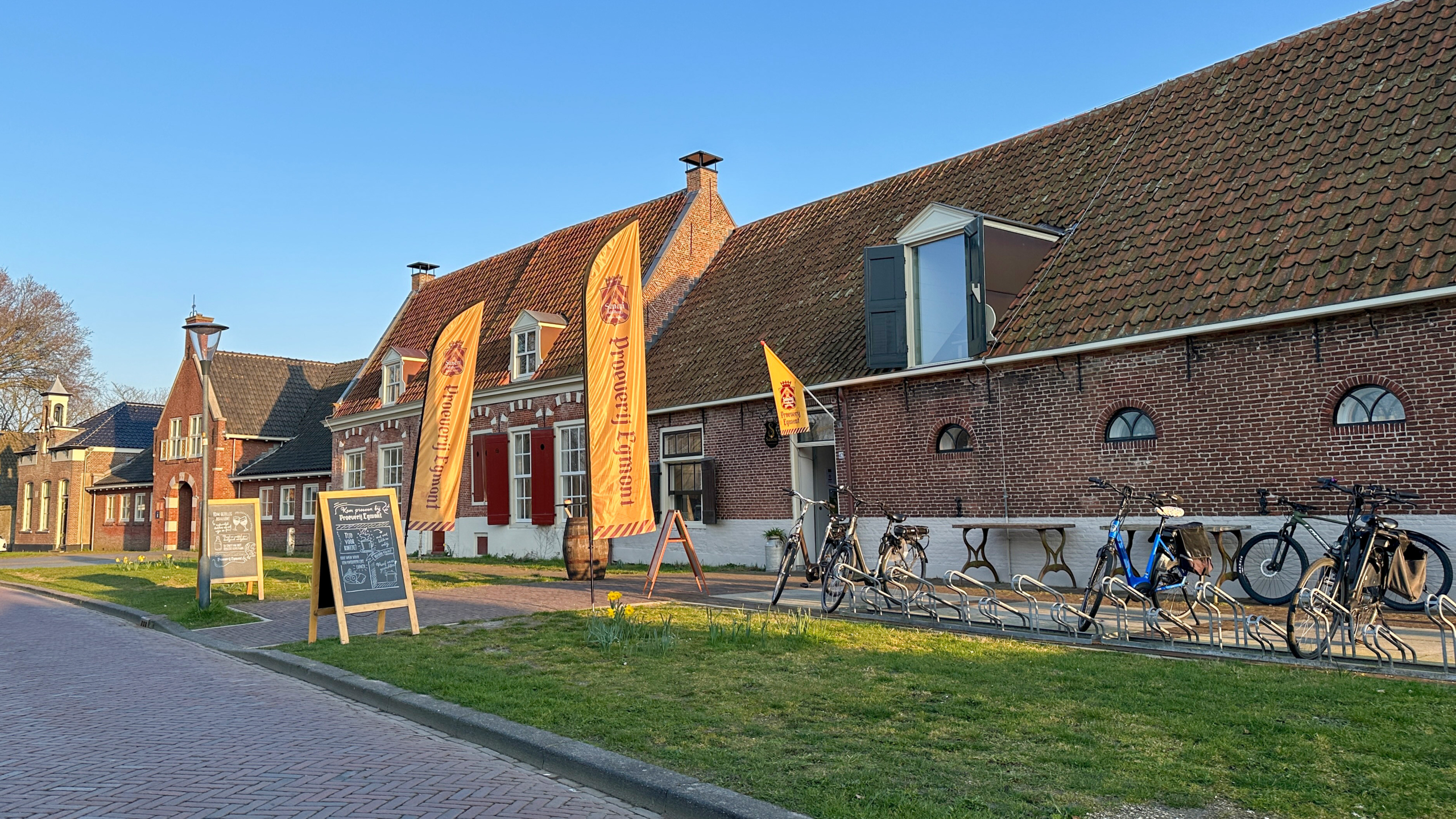 Rijtje historische bakstenen gebouwen met fietsenrekken en gele vlaggen van Brouwerij Emelisse langs de straat.