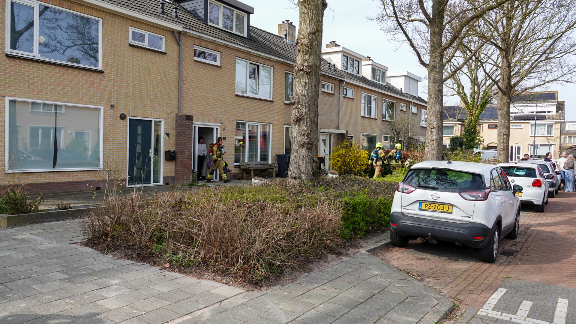 Brandweerlieden bij een rijtjeshuis, terwijl omstanders en geparkeerde auto's op straat zichtbaar zijn.