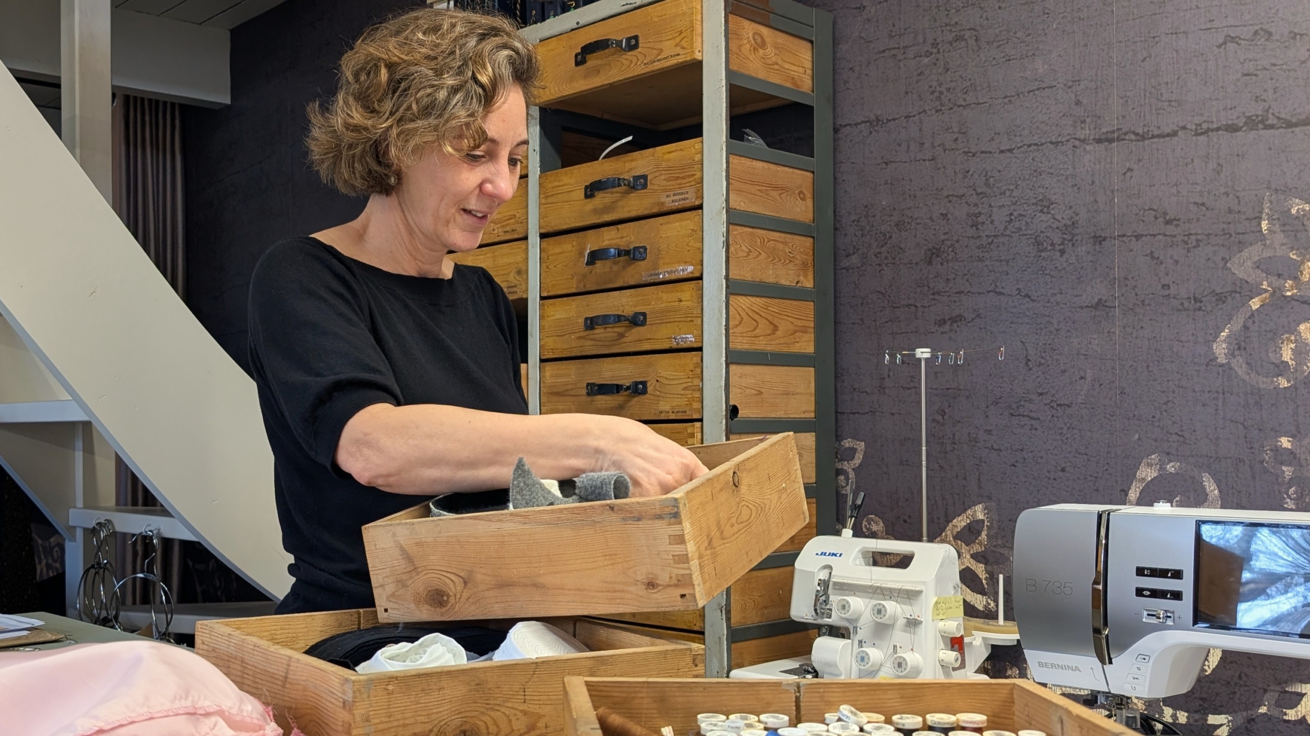 Vrouw kijkt in een houten lade vol stoffen en naaibenodigdheden, naast een naaimachine in een atelier.