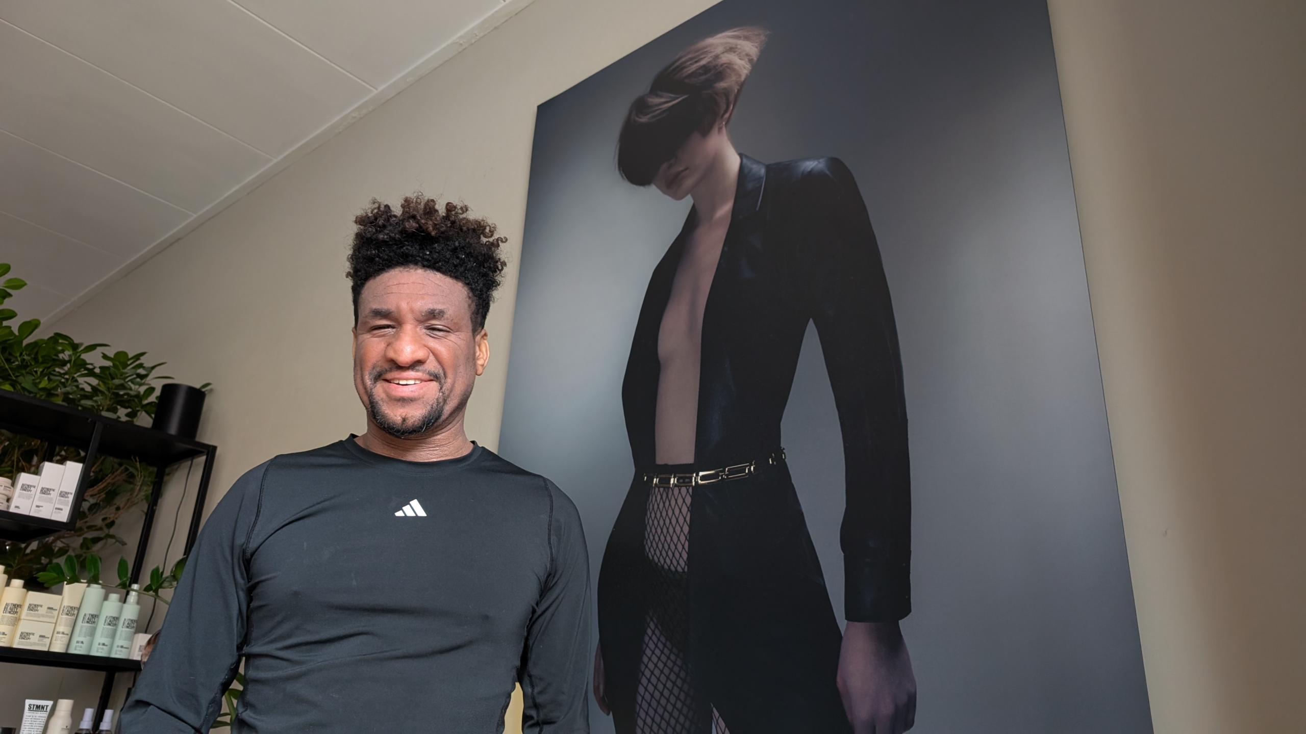 Man met krullend haar glimlacht voor een groot portret van een vrouw in een stoere outfit aan de muur.