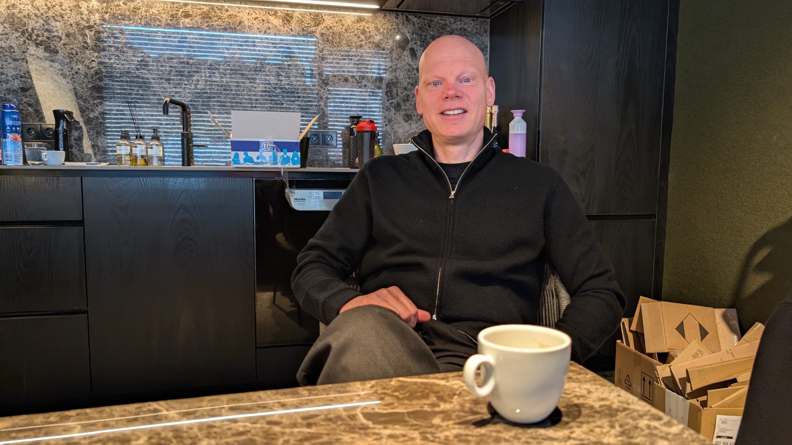 Man zit glimlachend aan tafel in een moderne keuken, met een kopje koffie voor zich en kartonnen dozen op de achtergrond.