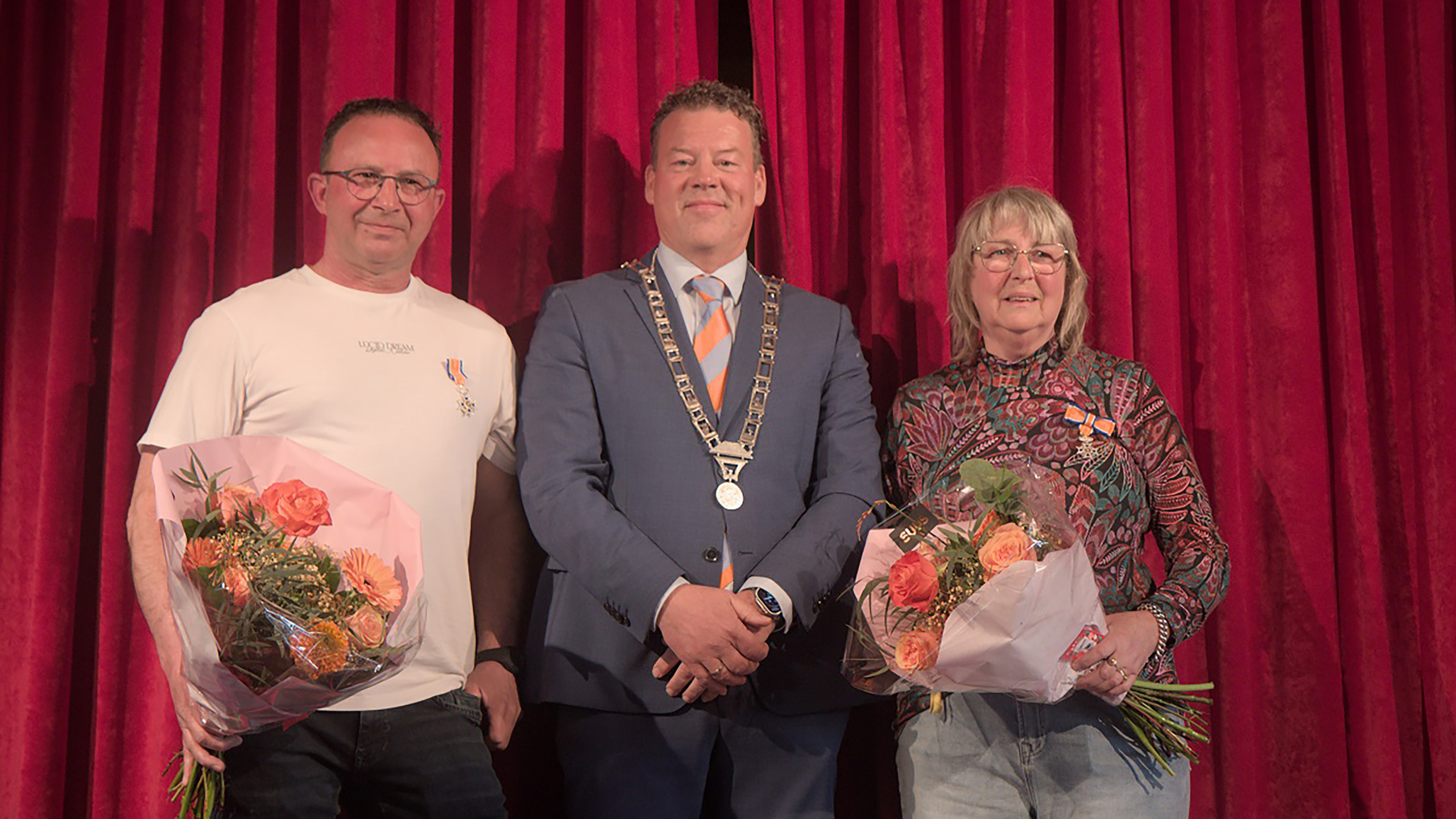 Twee mensen met bloemen staan naast een man in een blauw pak met ambtsketen voor een rode achtergrond.