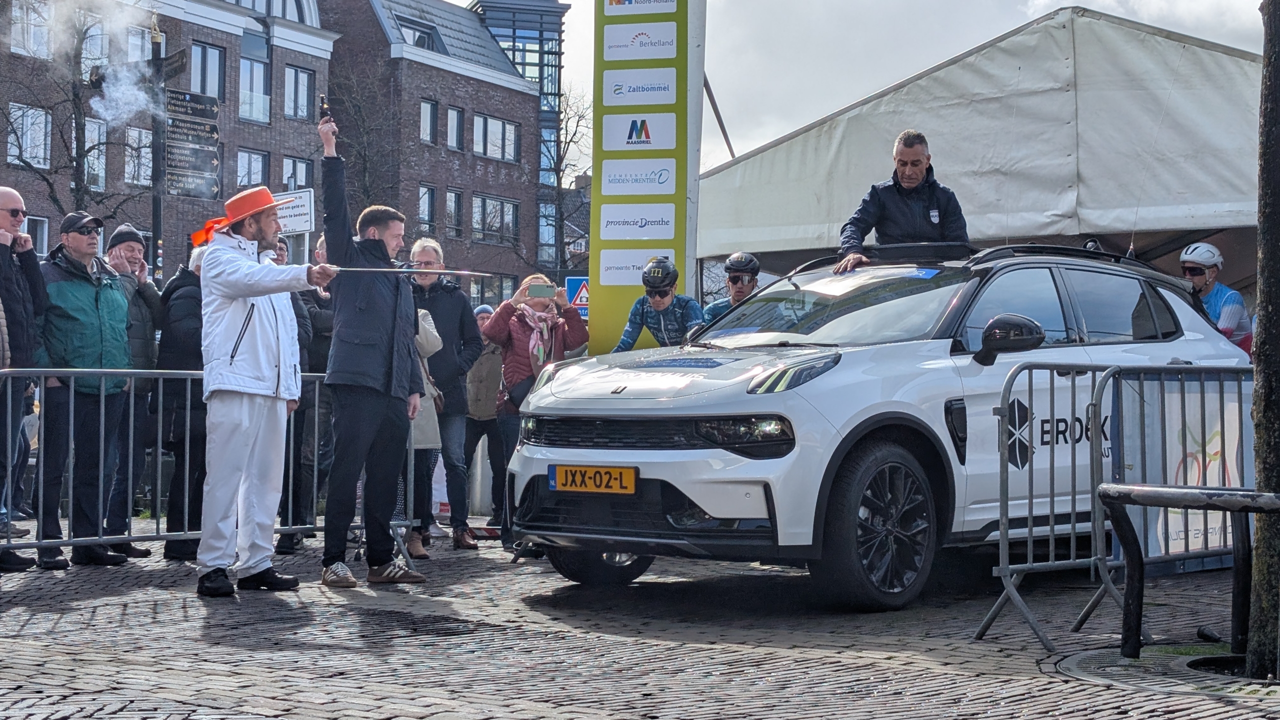 Mensen staan ​​rond een witte auto bij een evenement, met enkele personen in sportkleding en spandoeken op de achtergrond.