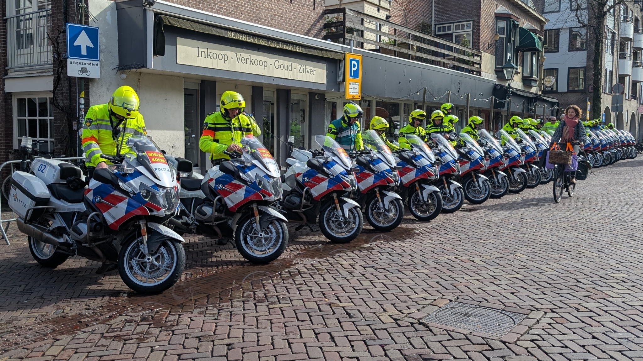 Een rij politieagenten op motoren gestationeerd naast een winkel, terwijl een vrouw op een fiets passeert.