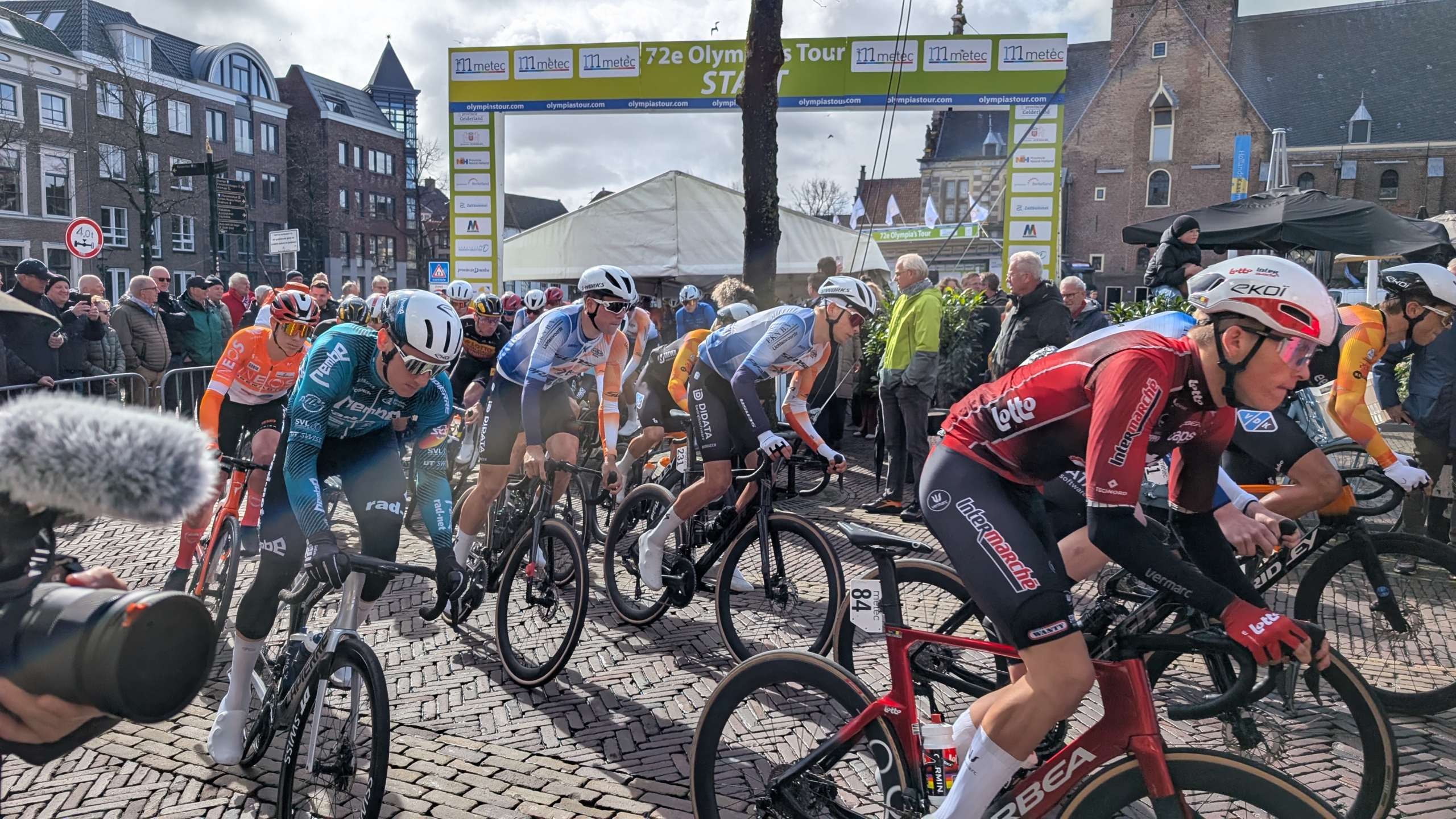 Wielrenners starten bij de 72e Olympia's Tour onder een boog, met toeschouwers op de achtergrond.