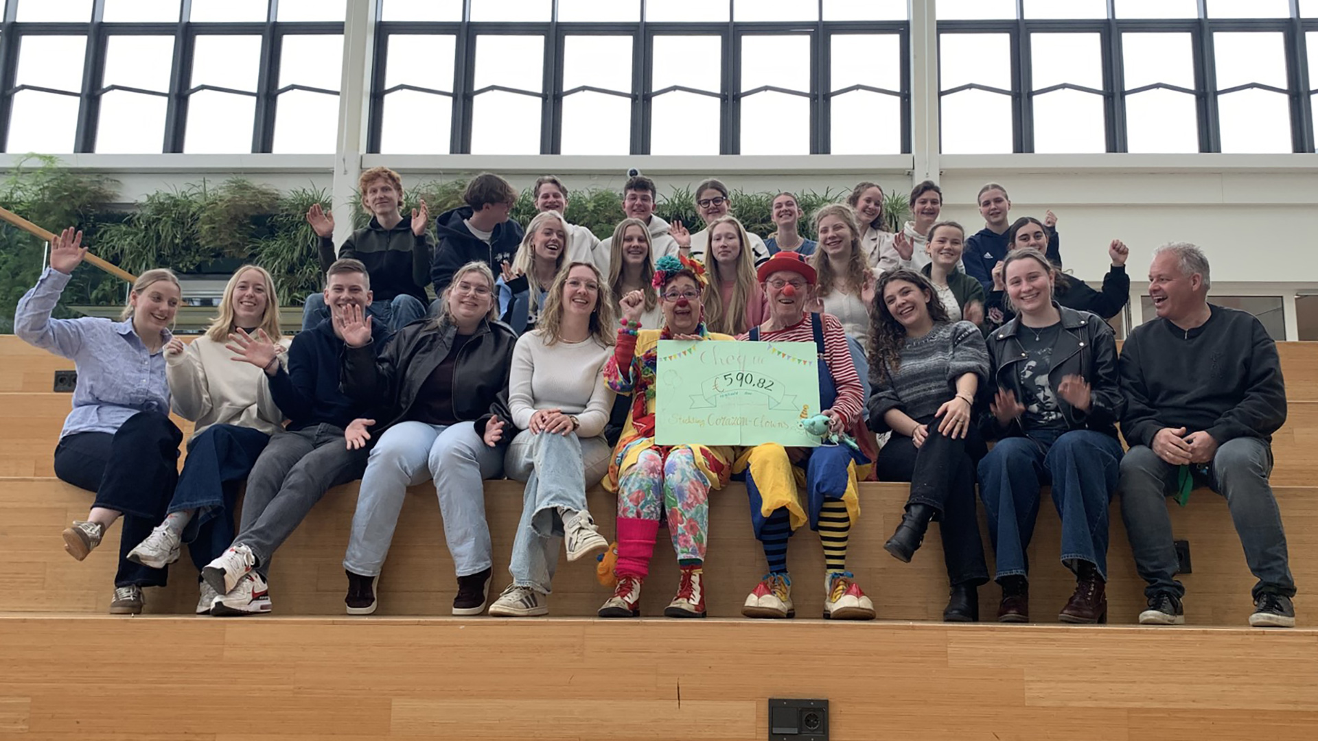Groep mensen, inclusief twee clowns met een cheque van 590,82 euro, zittend op trappen in een lichte ruimte, lachend en zwaaiend naar de camera.