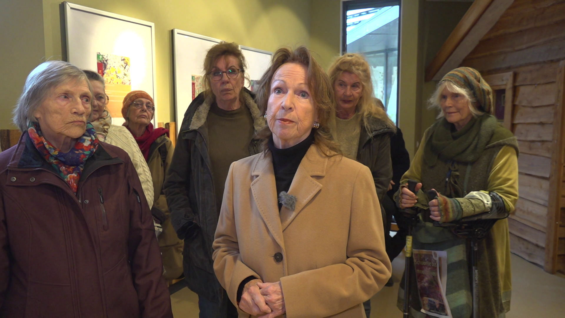Een groep oudere vrouwen staat in een ruimte met schilderijen aan de muur, de vrouw in het midden draagt een beige jas.