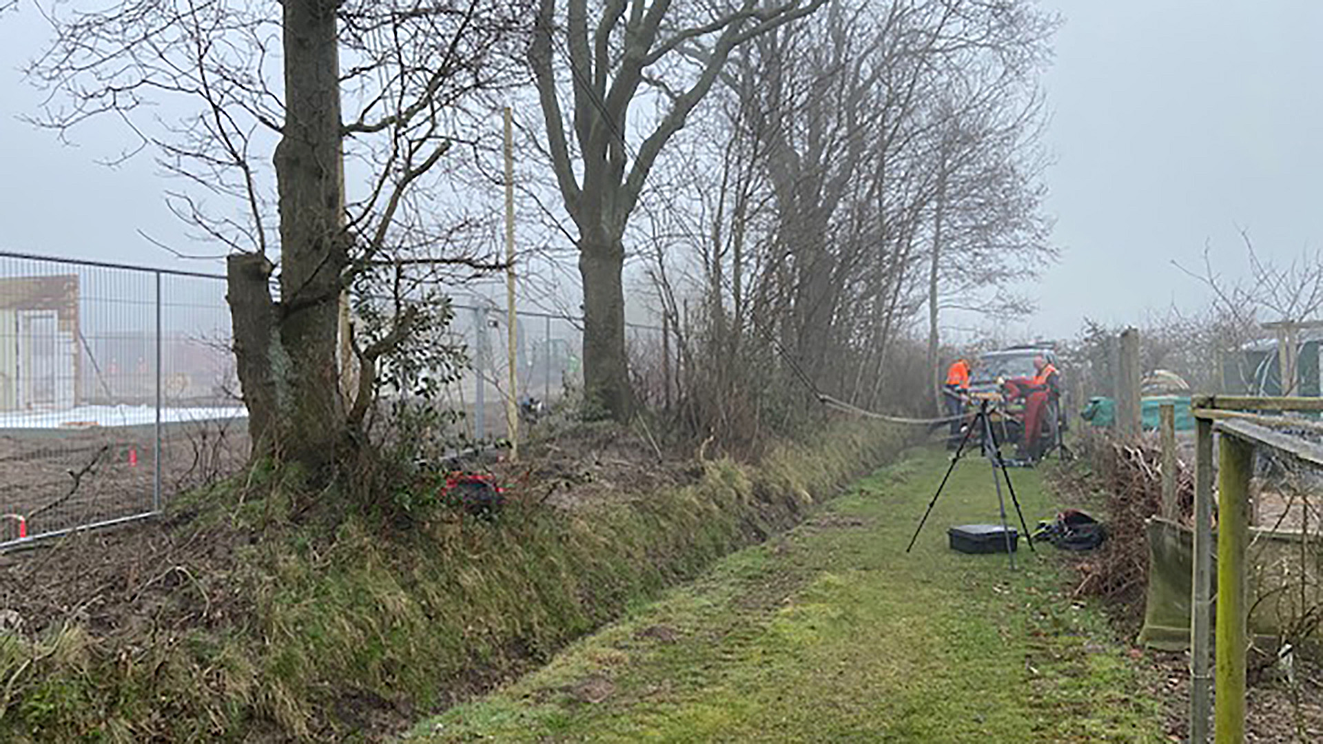 Twee mensen in oranje werkjassen werken naast een hek aan de rand van een grasveld met bomen en gereedschap.