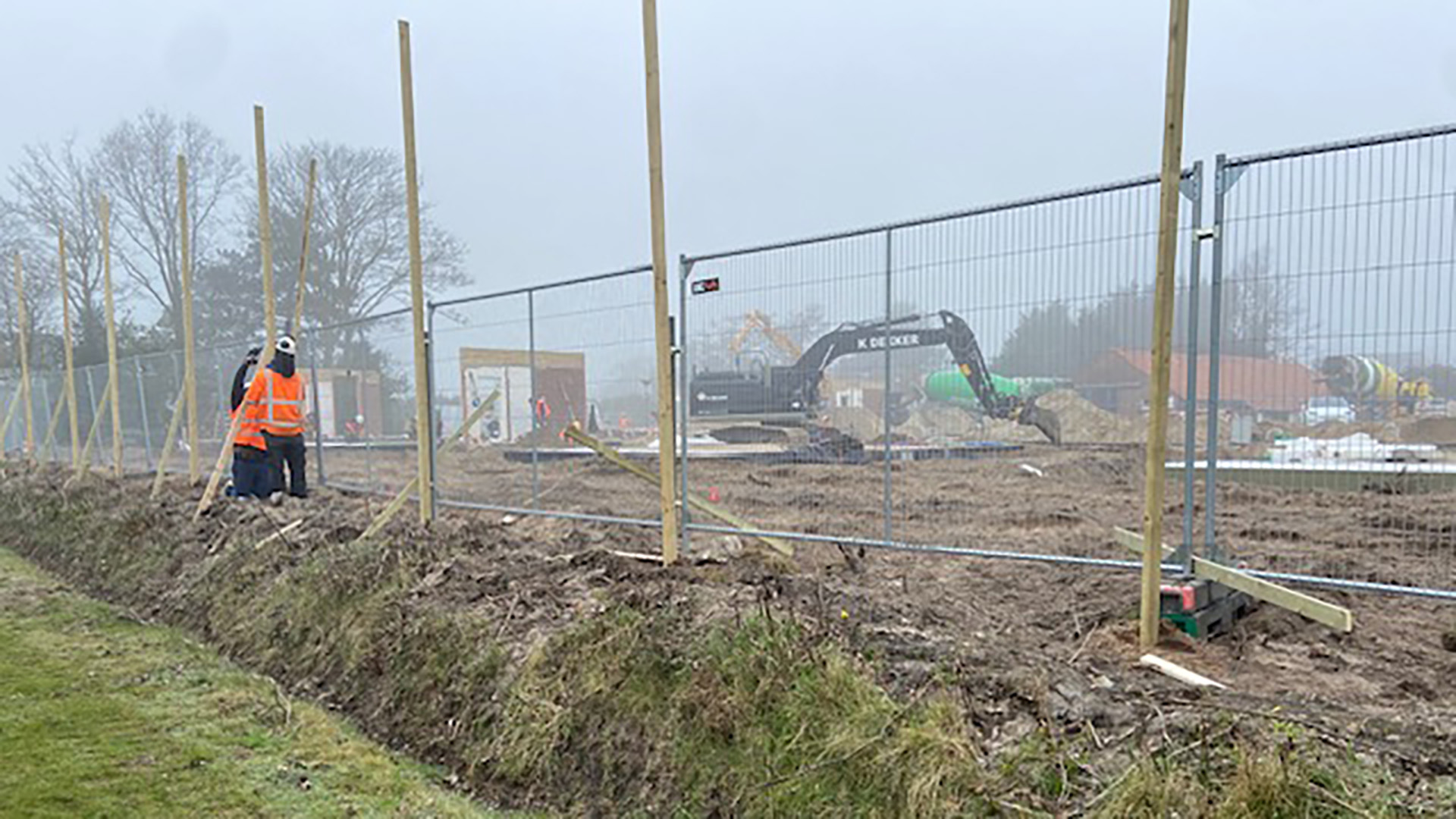 Bouwterrein met een graafmachine en een persoon in een oranje veiligheidsvest, omgeven door hekken en houten palen.
