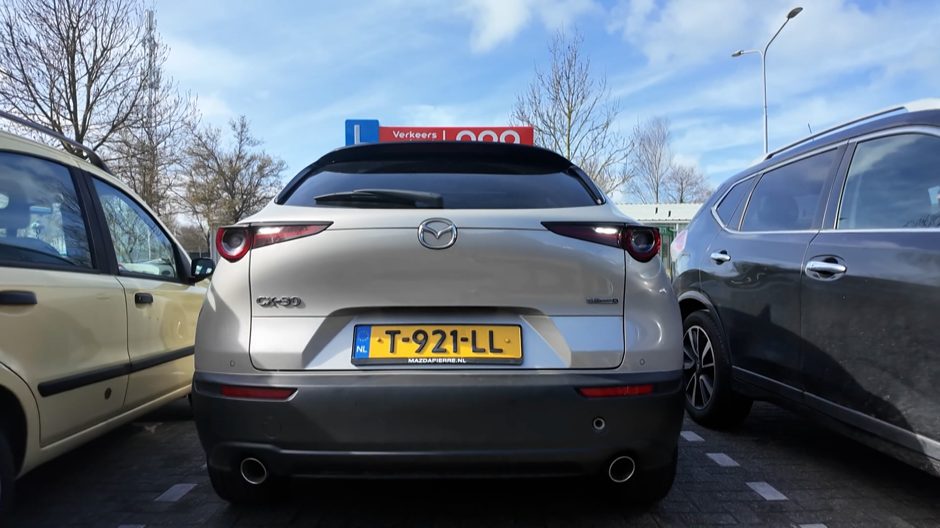 Grijze Mazda CX-30 auto, geparkeerd tussen twee andere auto's, met Nederlandse kentekenplaat T-921-LL.