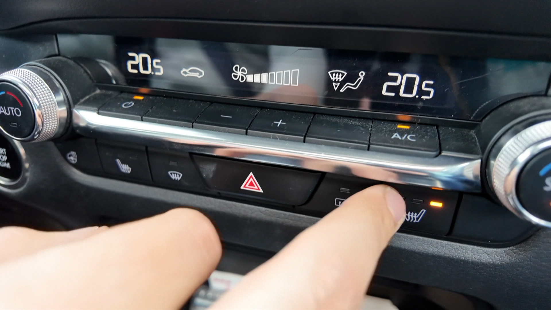 Vinger drukt op de stoelverwarmingsknop in de auto, met buitenluchtinstelling op 20,5 graden en ventilatorinstellingen zichtbaar.