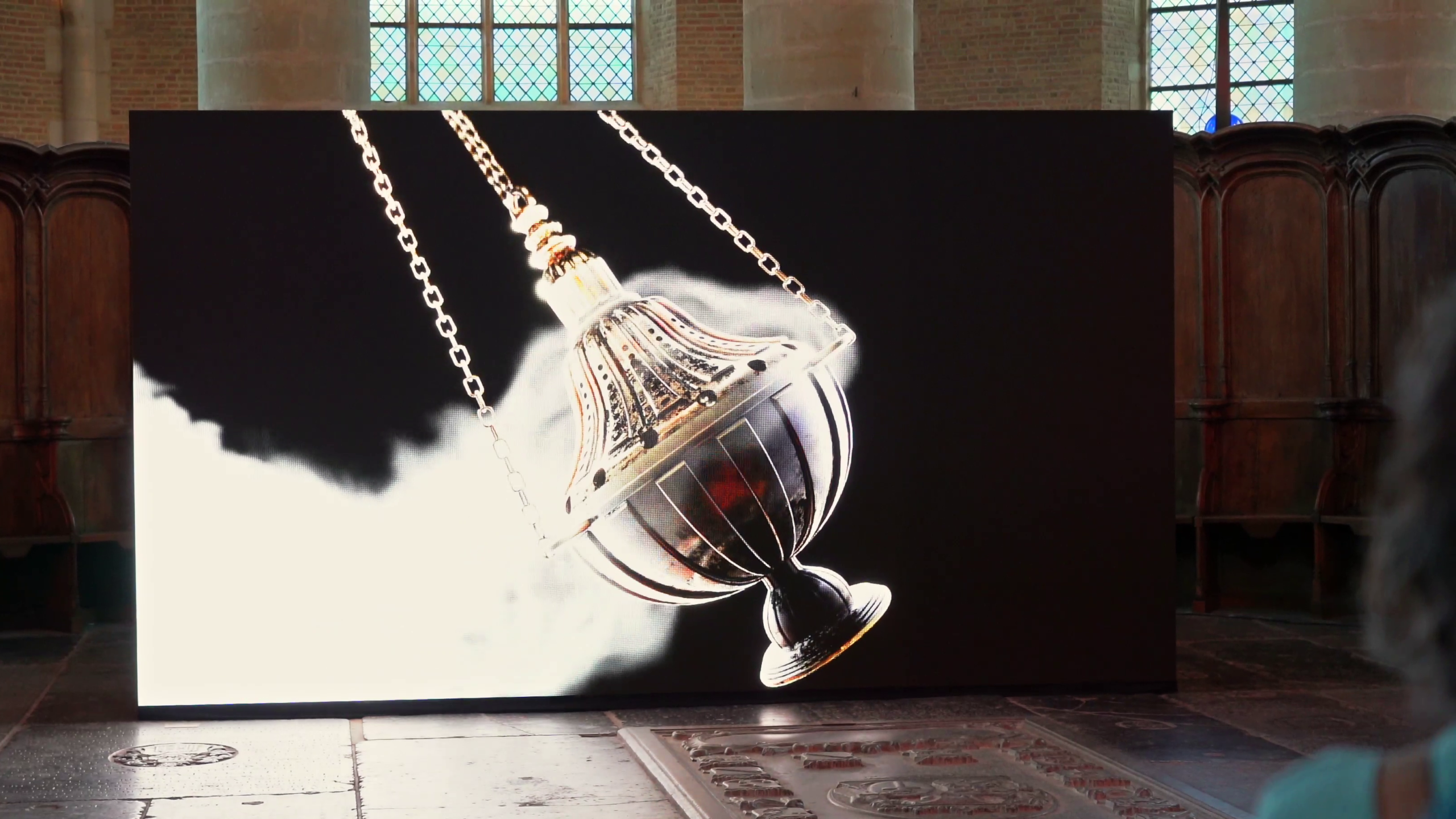 Een groot scherm toont een wierookvat aan kettingen, met rook tegen een zwarte achtergrond in een kerkelijke omgeving.