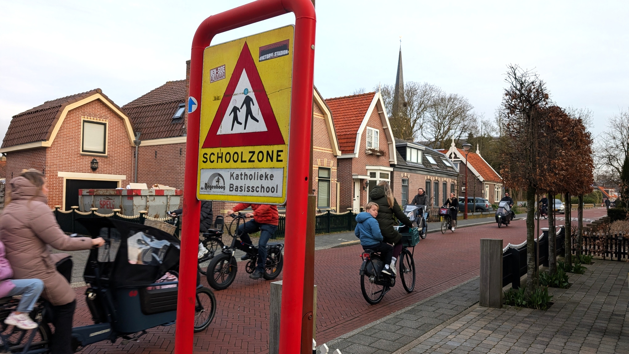 Verkeersbord met 'Schoolzone' en 'Katholieke Basisschool'; fietsende volwassenen en kinderen in een woonwijk.