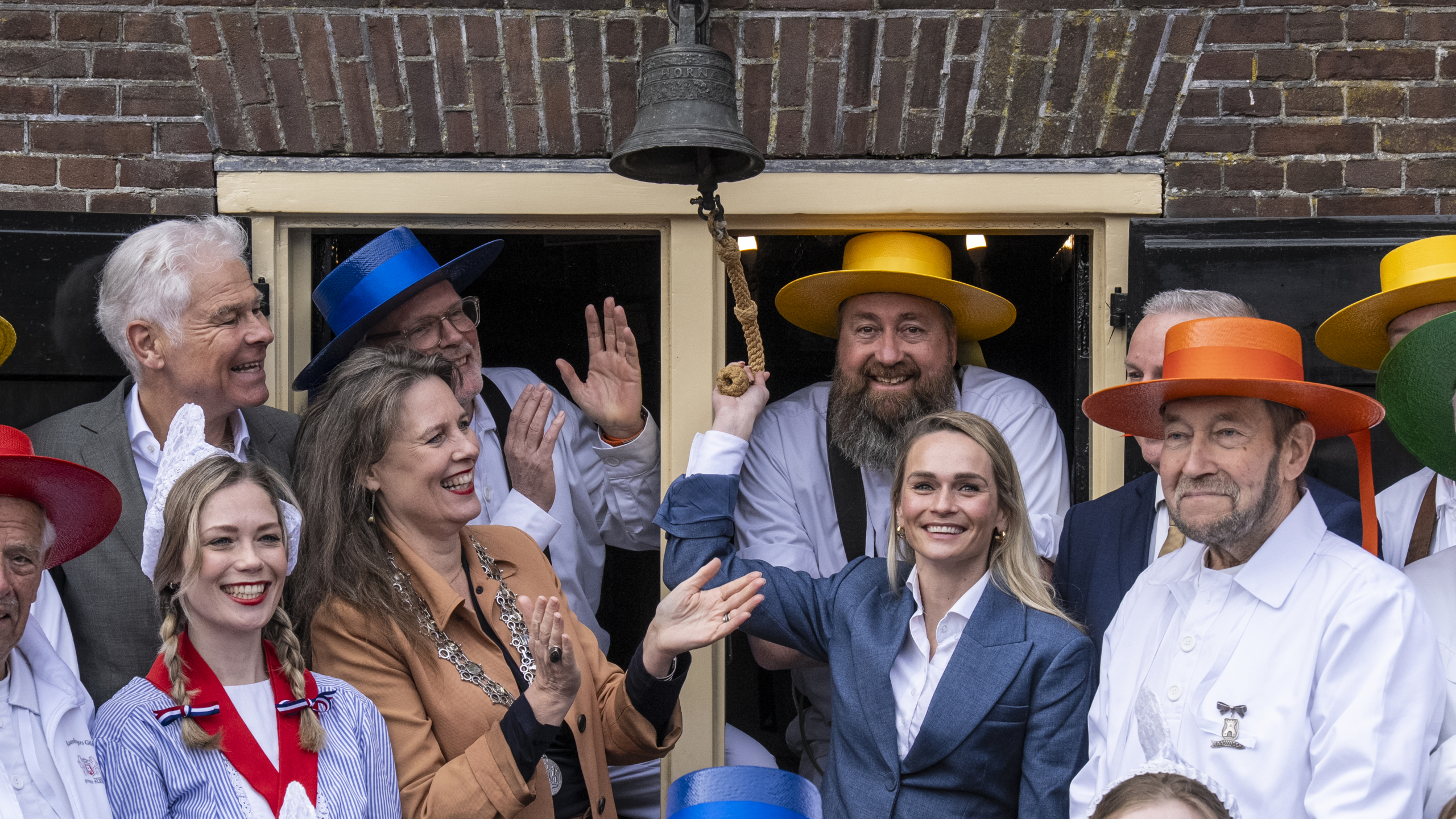 Een groep mensen in traditionele klederdracht poseert lachend voor een gebouw, terwijl iemand een bel luidt.
