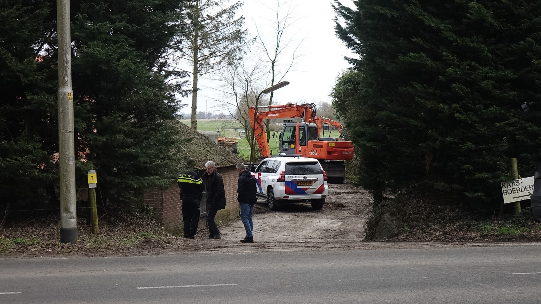 Politieagenten en een politiewagen staan bij een bouwplaats met een graafmachine, omgeven door bomen.