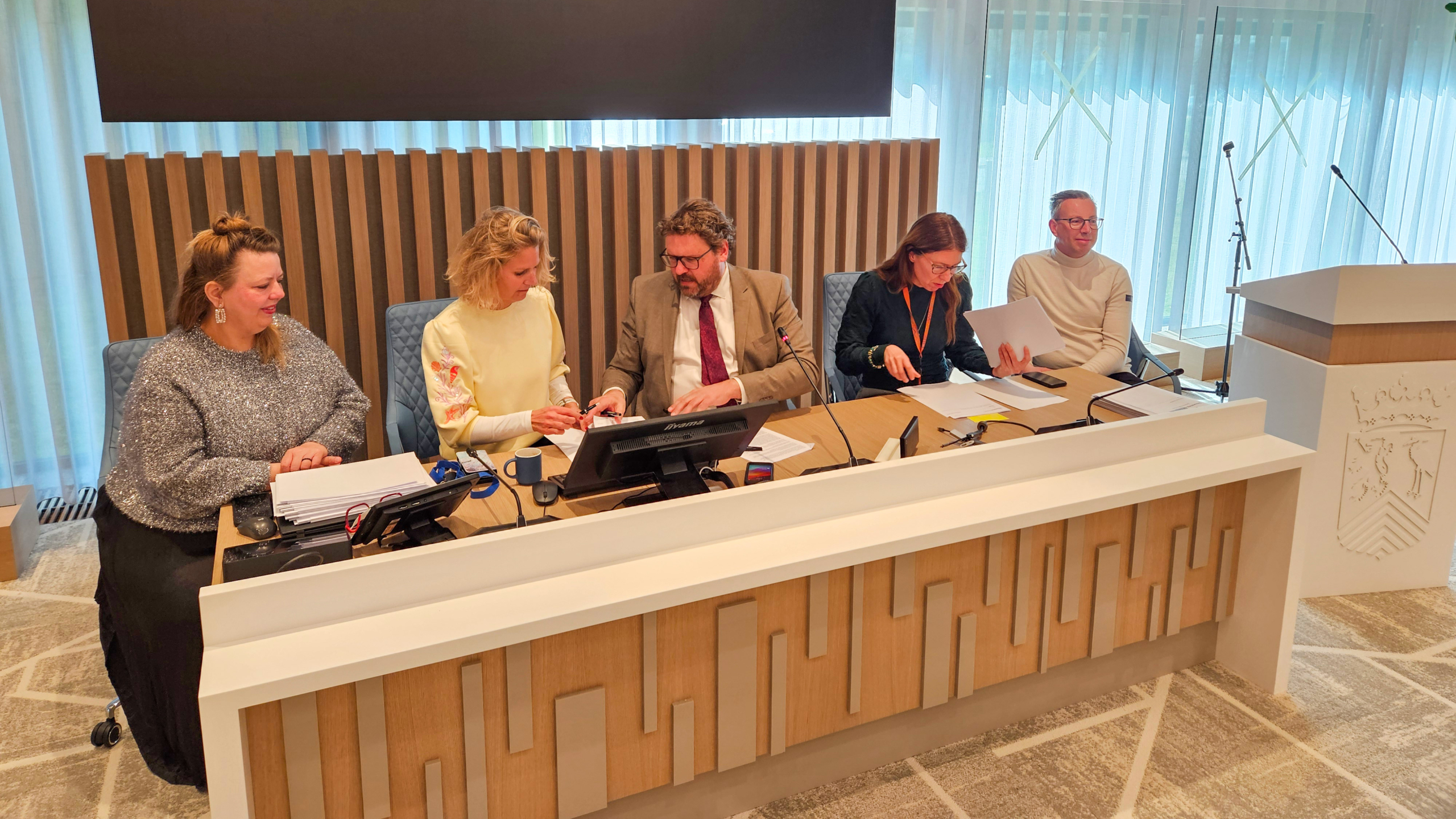 Vijf mensen zitten achter een bureau in een vergaderruimte, met documenten en microfoons voor hen.