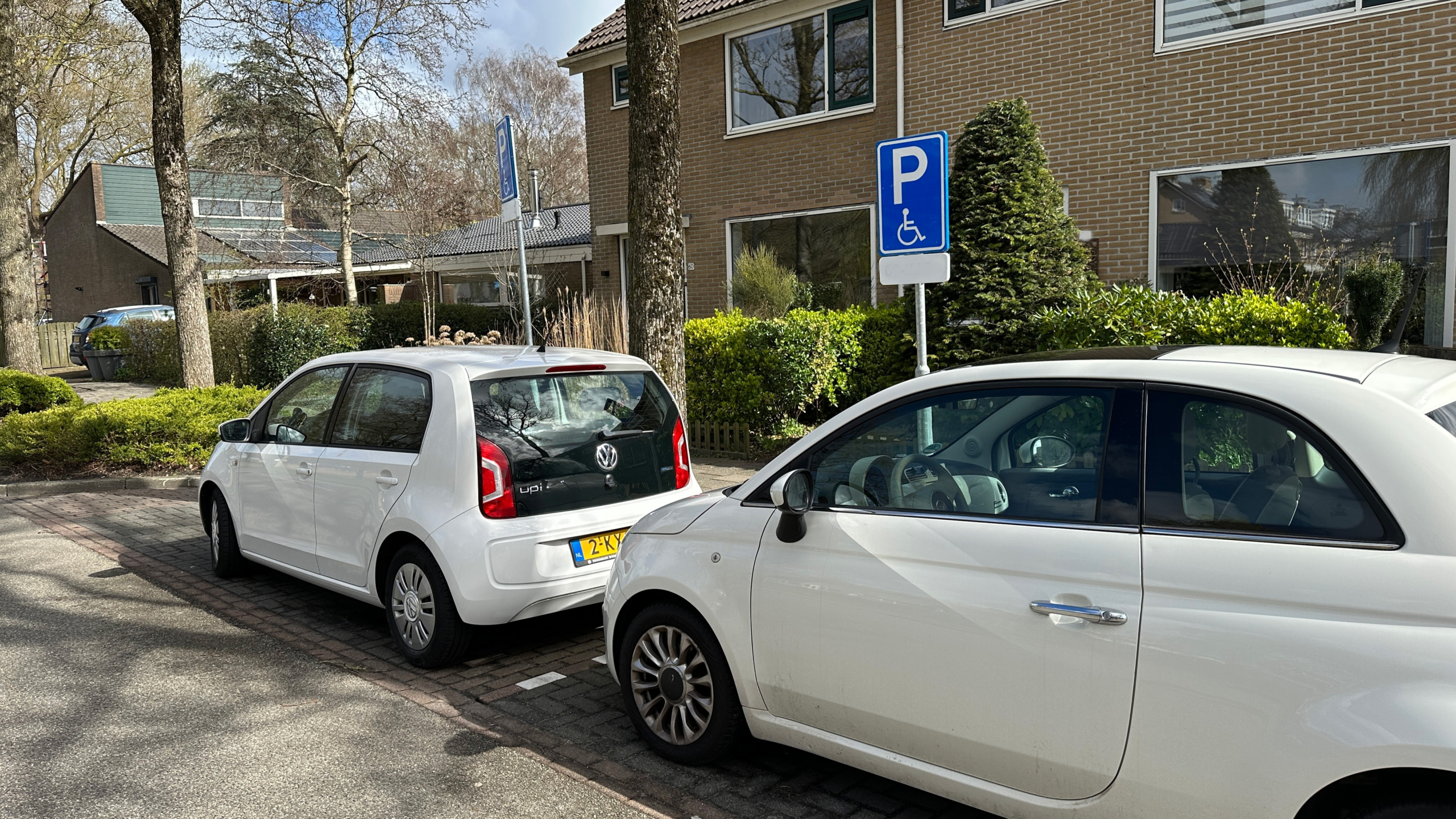 Twee auto's geparkeerd bij een gehandicaptenparkeerplaats voor een bakstenen huis met groene struiken.
