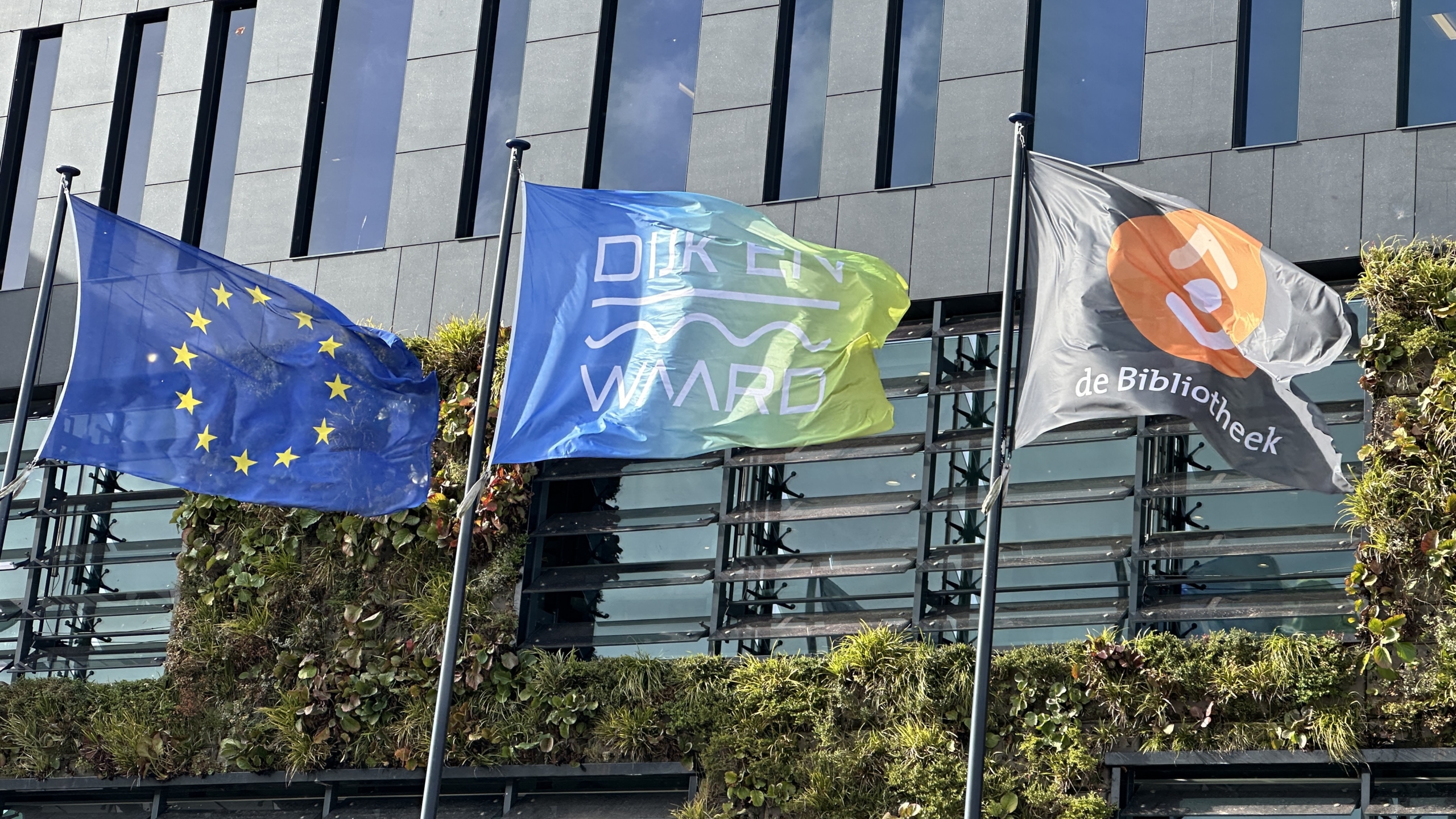 Drie vlaggen wapperen voor een gebouw met verticale tuinen: de EU-vlag, "Dijk en Waard," en de Nederlandse bibliotheekvlag.