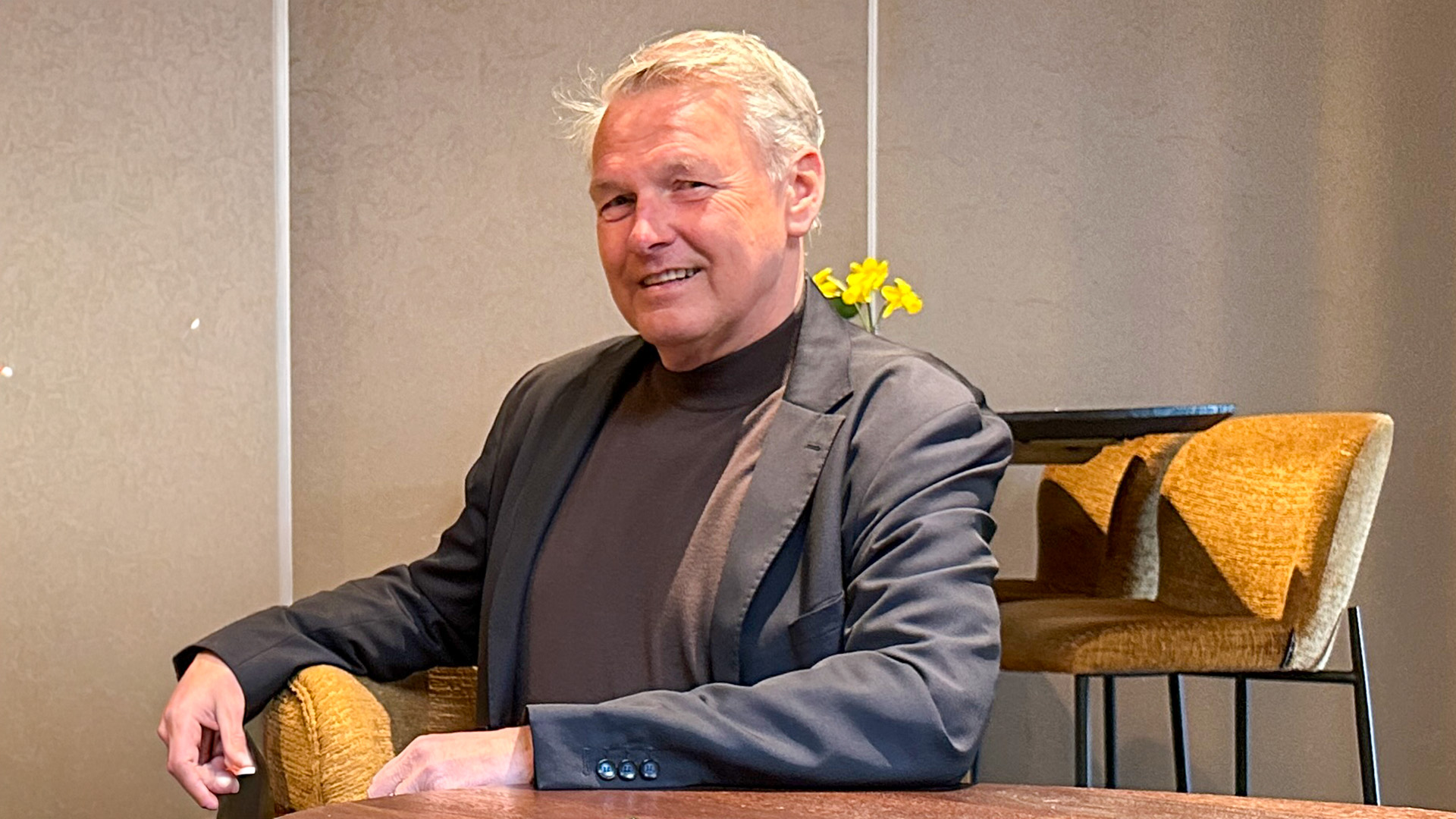 Een man in een donkere trui en jasje zit aan een tafel, met gele bloemen op de achtergrond.