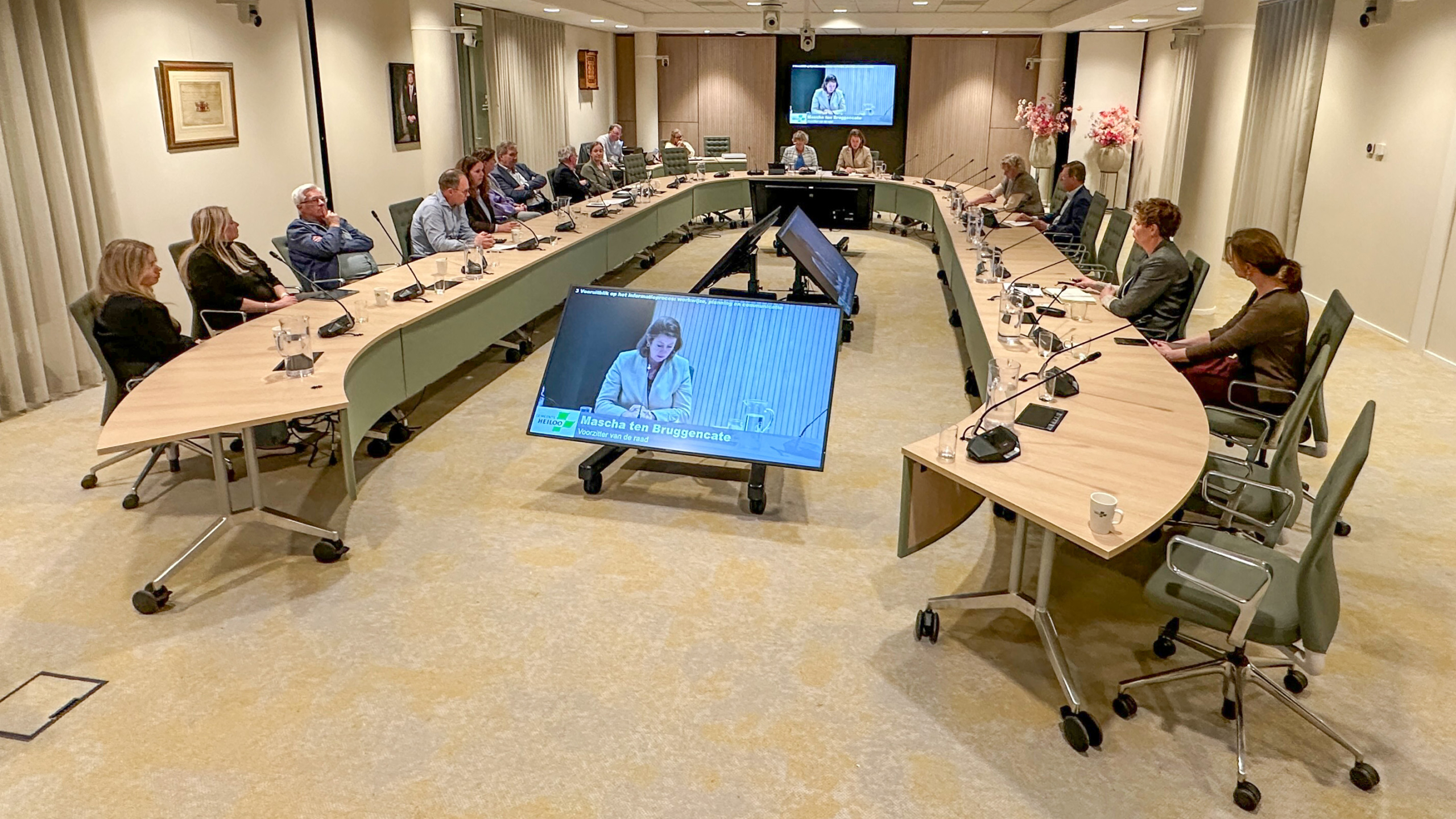 Mensen zitten in een vergaderruimte rondom een U-vormige tafel met microfoons; een scherm toont een vrouw die spreekt.