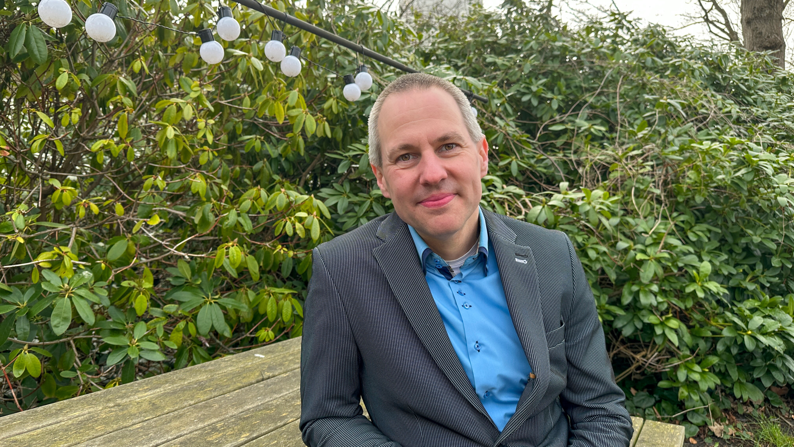 Man in blauw overhemd en geruit jasje zit aan houten tafel in een tuin met groene struiken en hangende lampjes.