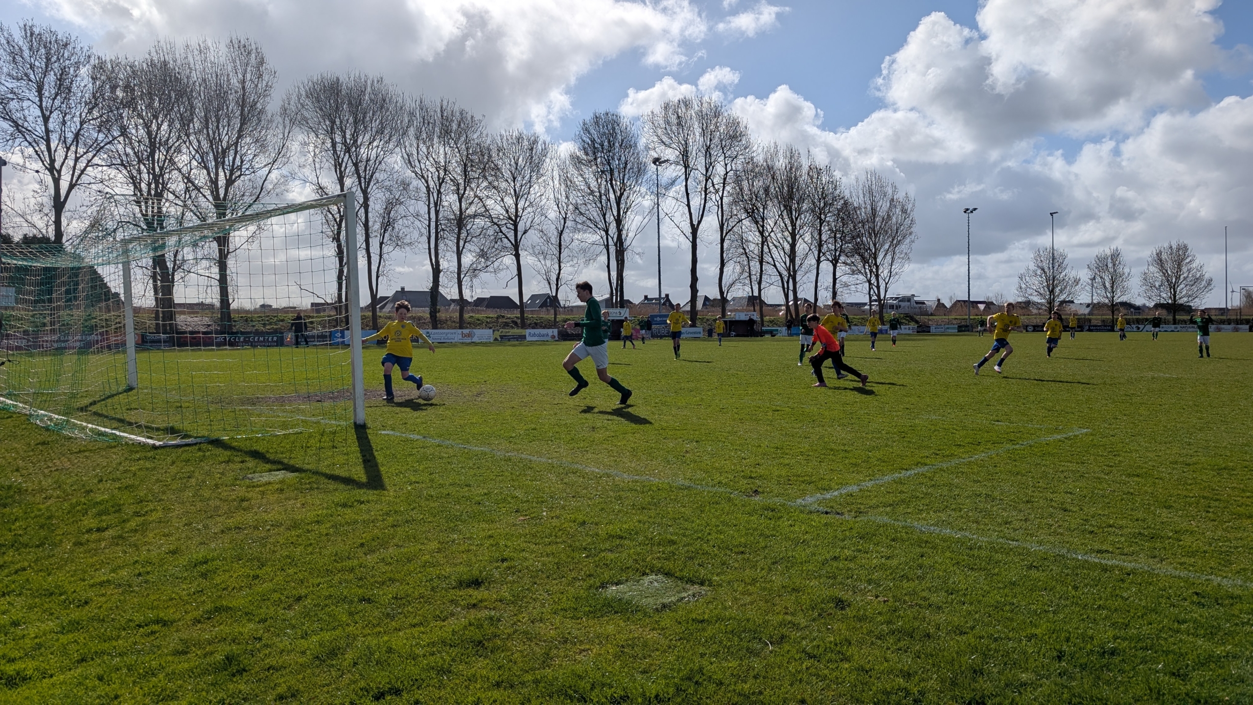 Voetballers in actie op een voetbalveld, een speler in geel probeert de bal te scoren, terwijl een tegenstander nadert.