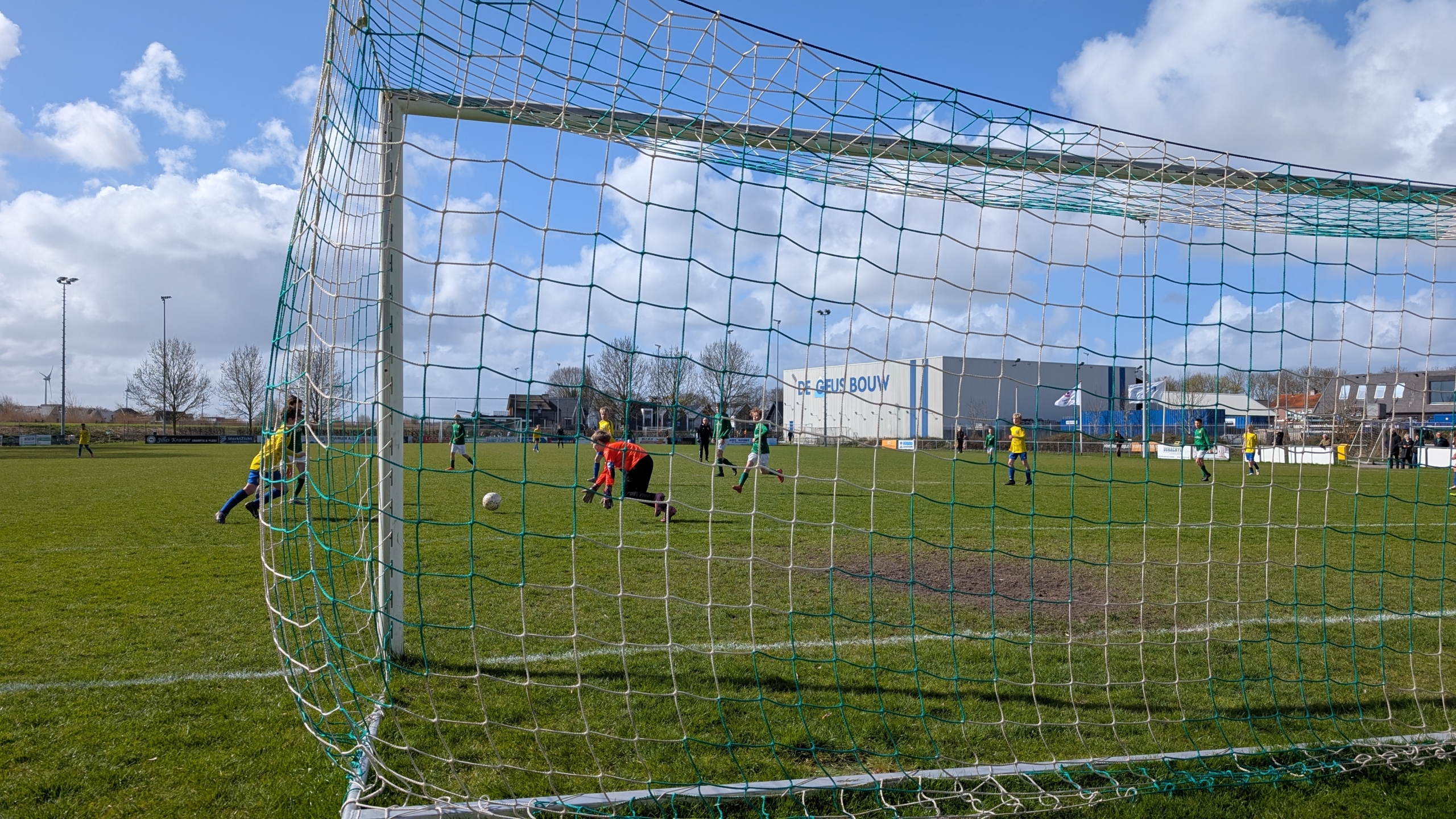 Voetbalwedstrijd met een keeper in rood die een bal probeert te stoppen vanuit het perspectief van het doel, met spelers in geel en groen.