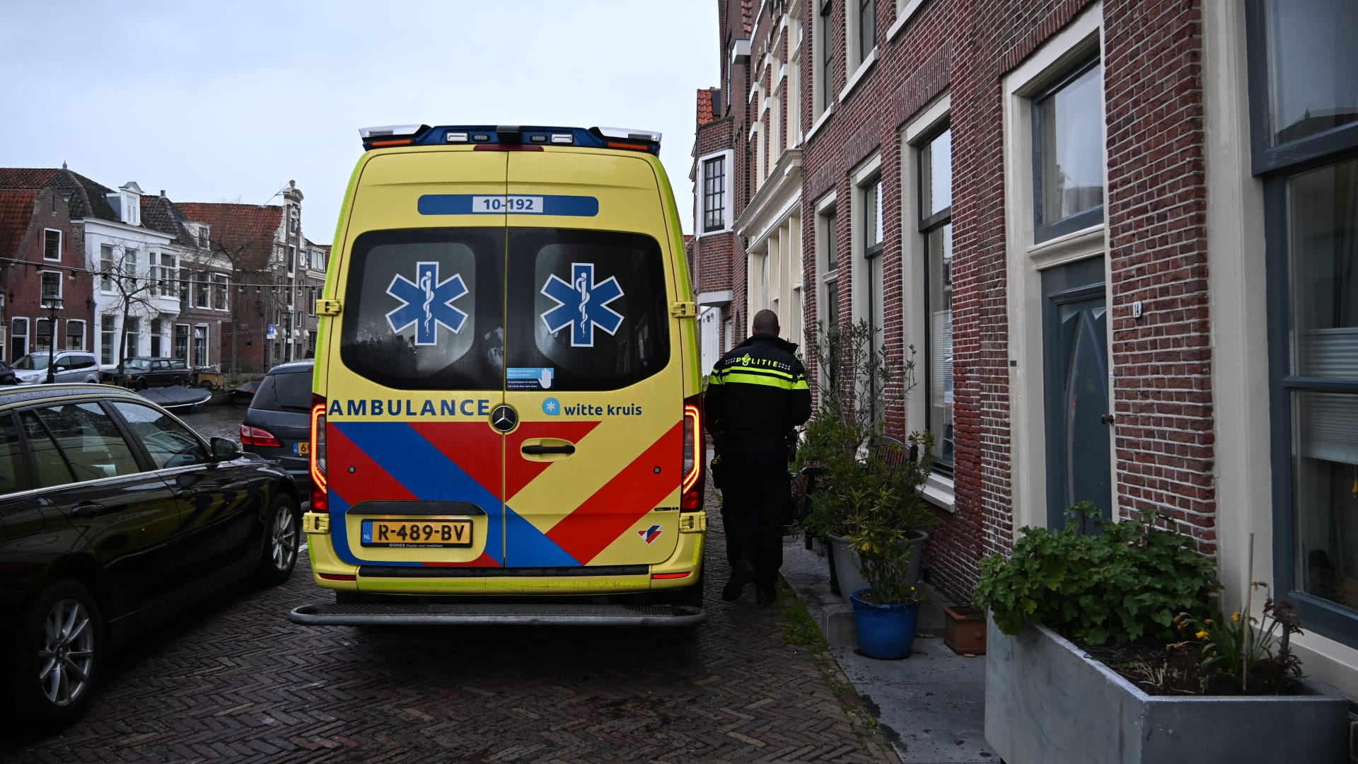 Een ambulance en een politieagent bij een bakstenen gebouw in een smalle straat, met geparkeerde auto's en grachtenhuizen op de achtergrond.