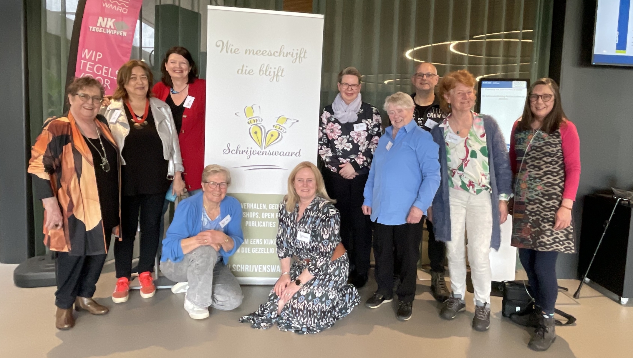 Groep mensen poseert voor een bord met de tekst "Wie meeschrijft die blijft, Schrijvenswaard," in een interieur.