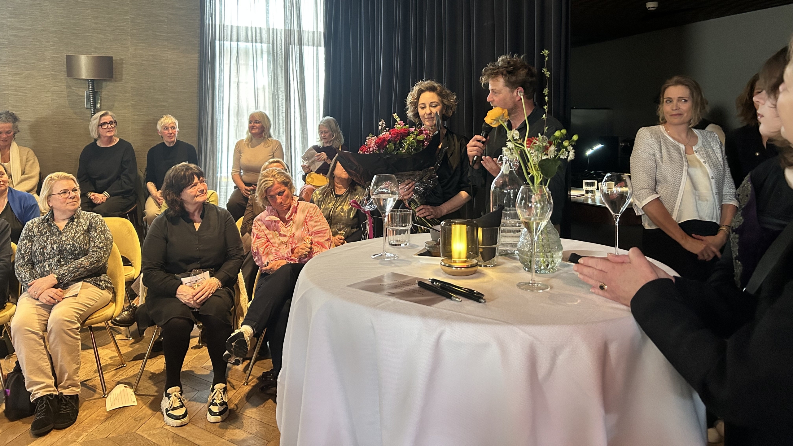 Mensen zitten en staan in een kamer, een persoon houdt een boeket bloemen en een microfoon vast, met mensen die toekijken.