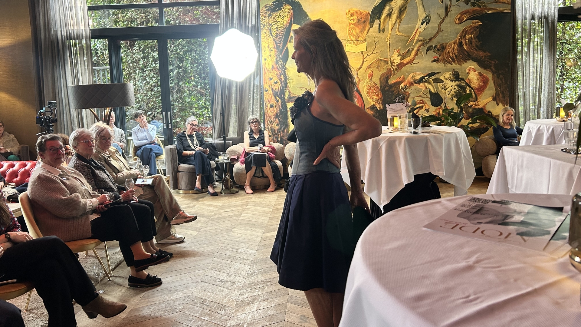 Vrouw in blauwe jurk presenteert iets aan een zittend publiek in een elegante ruimte met grote muurschildering.