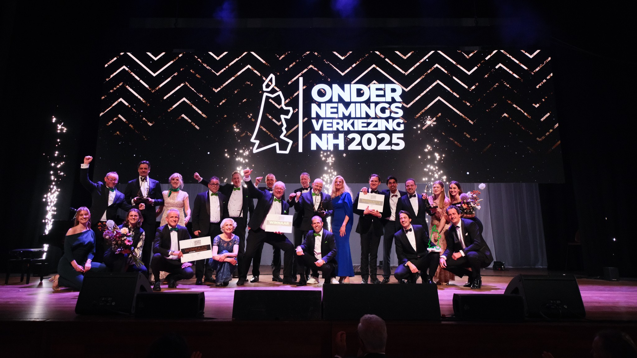 Groep mensen in avondkleding viert een prijsuitreiking op een podium met een groot scherm dat "Ondernemingsverkiezing NH 2025" toont.