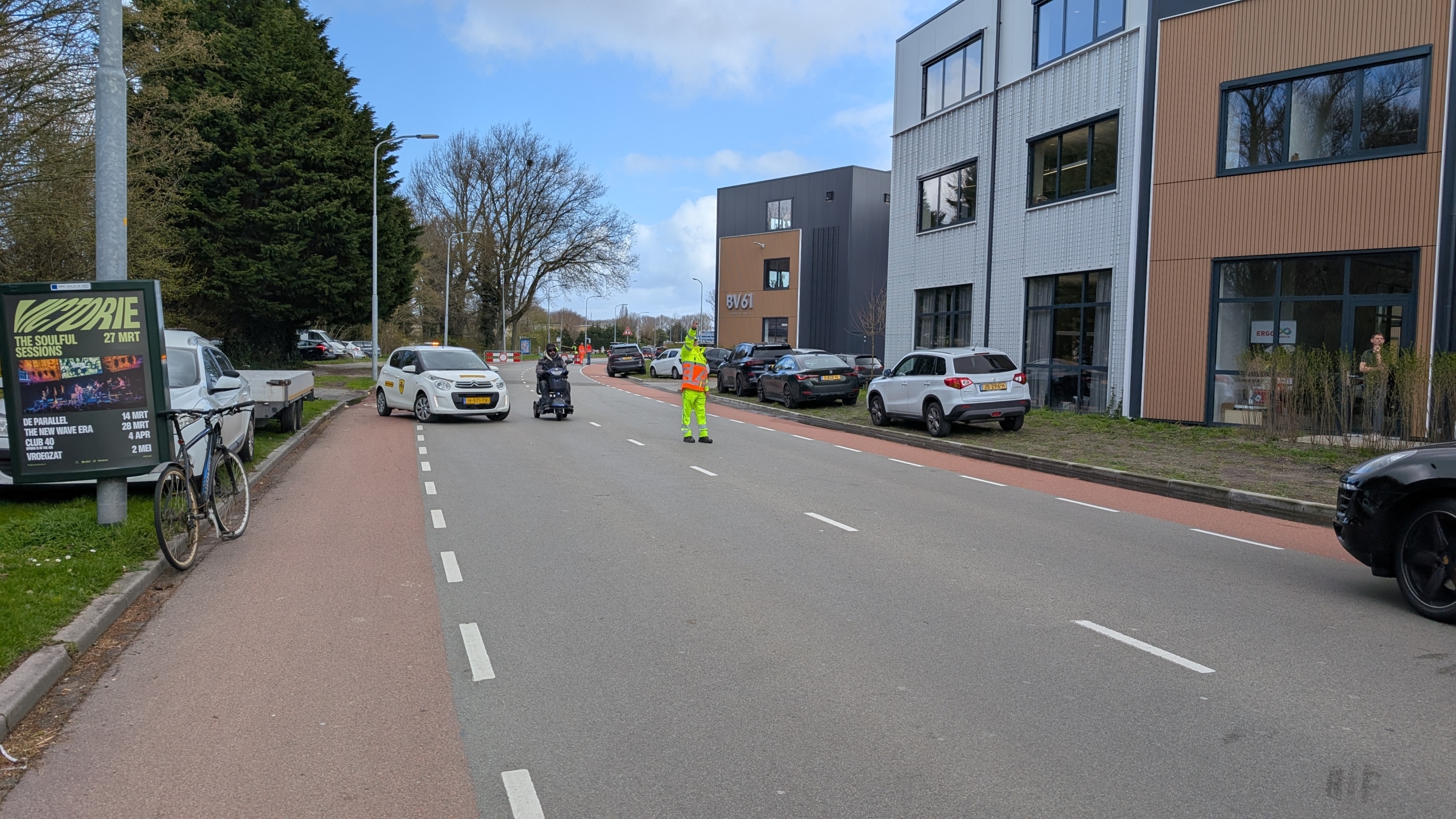 Een verkeersregelaar in een gele outfit staat op de weg en helpt een scootmobiel oversteken, met auto's op de achtergrond.