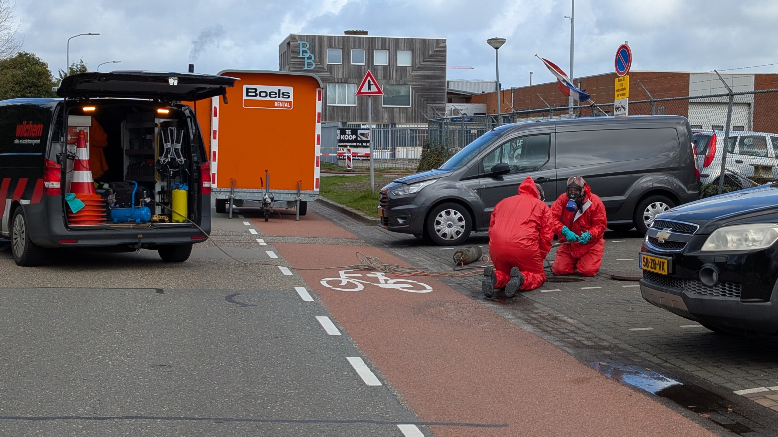 Twee mensen in rode veiligheidskleding werken naast een geparkeerde bestelwagen en open bus, met gereedschap op de grond.